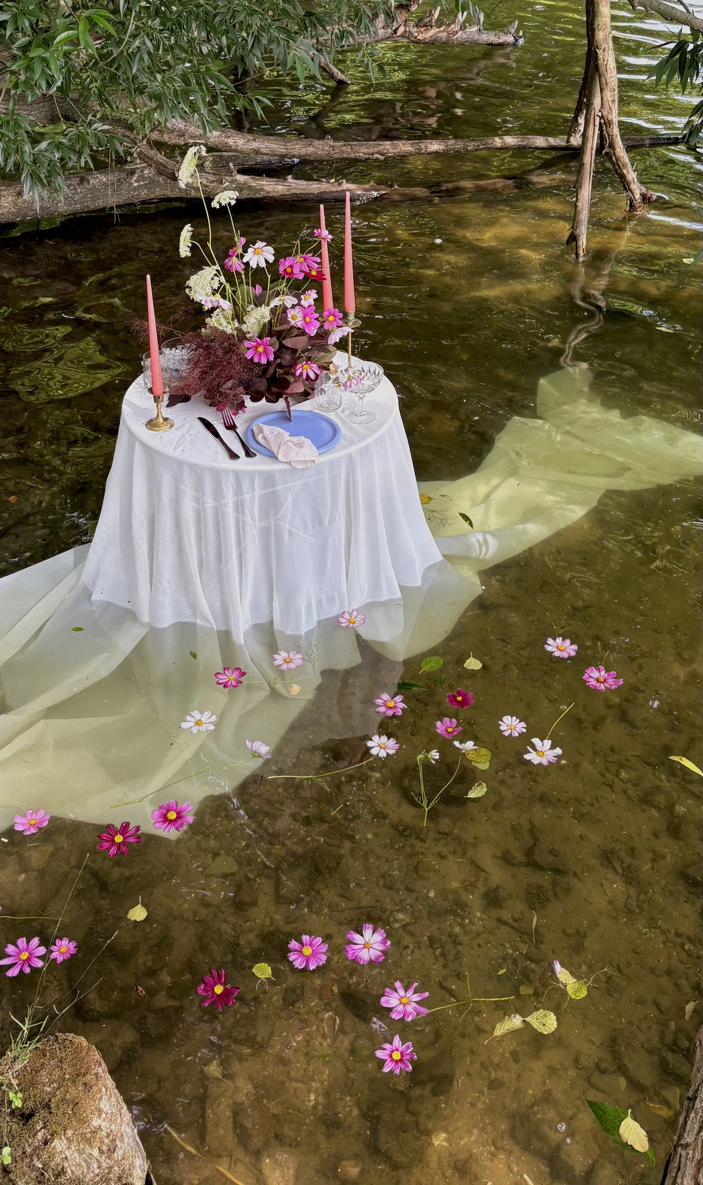 Elegant white table setting with pink candles and flowers beside a tree-lined pond, with floating flowers on the water