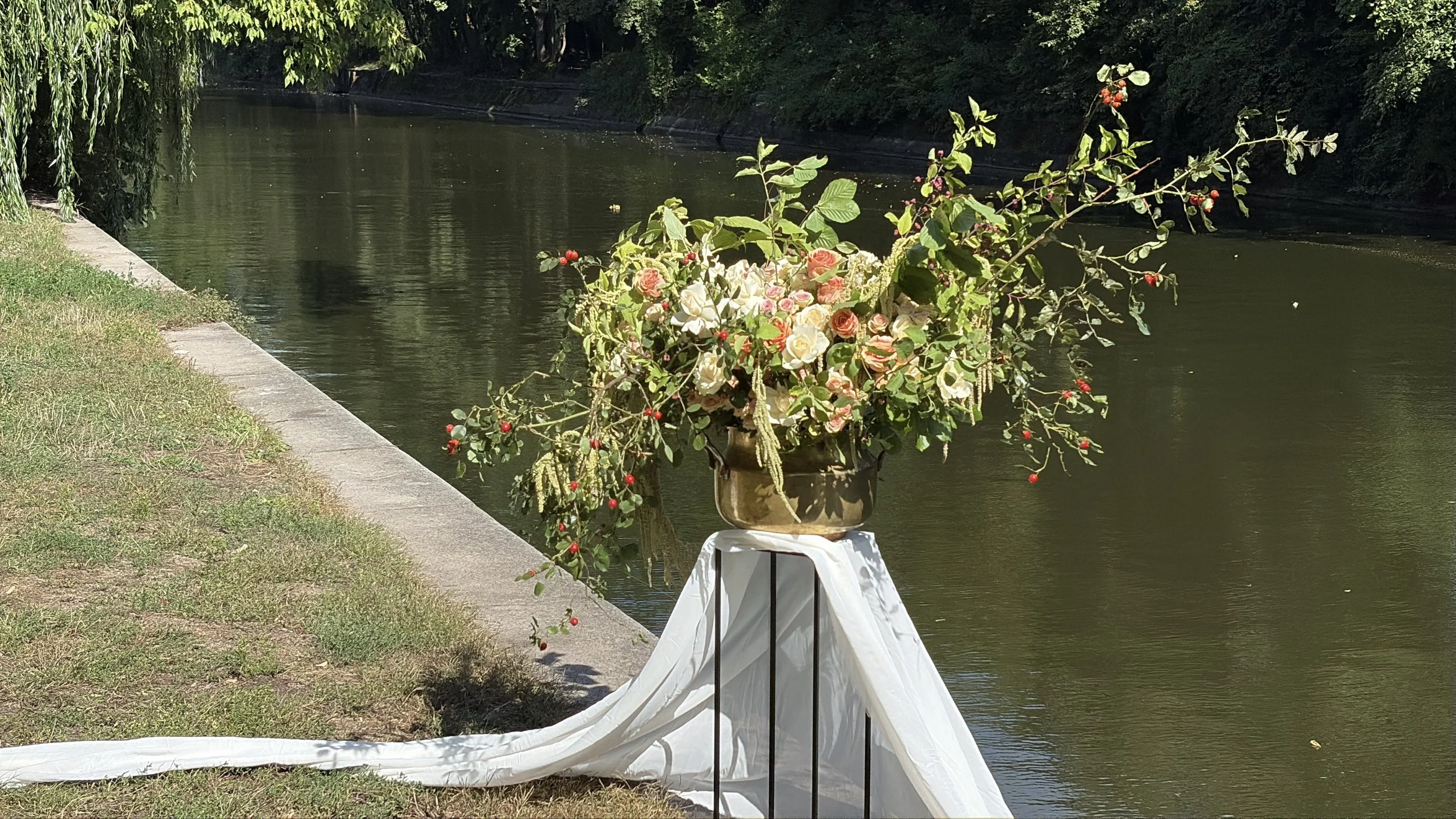 Floral arrangement of roses and red berries on a draped pedestal beside a tree-lined pond
