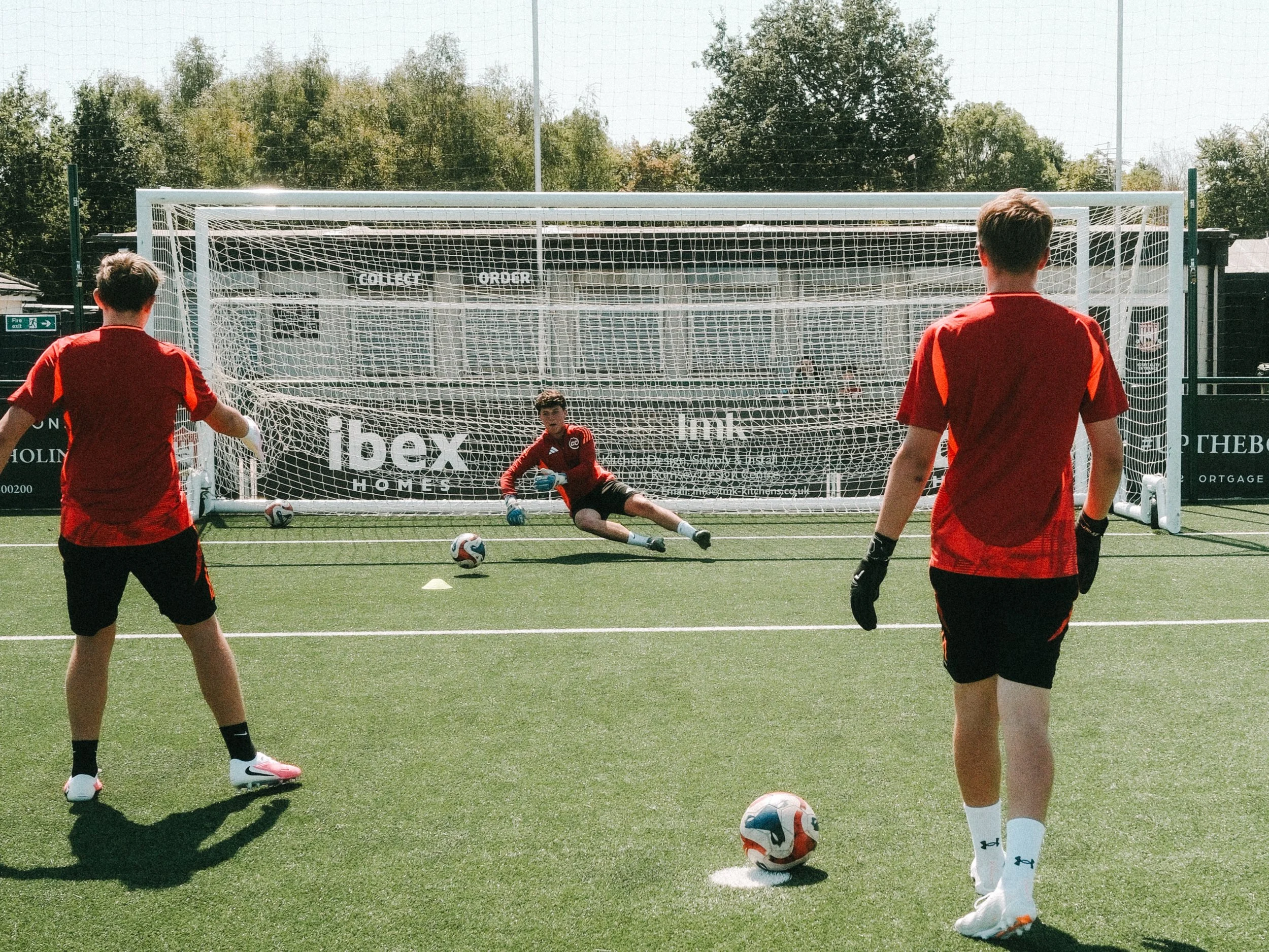 Young soccer players in red jerseys are practicing penalty kicks on an outdoor field. One player is about to take a shot, while another is preparing to kick, and a goalkeeper in red is sitting down in front of the goal post.