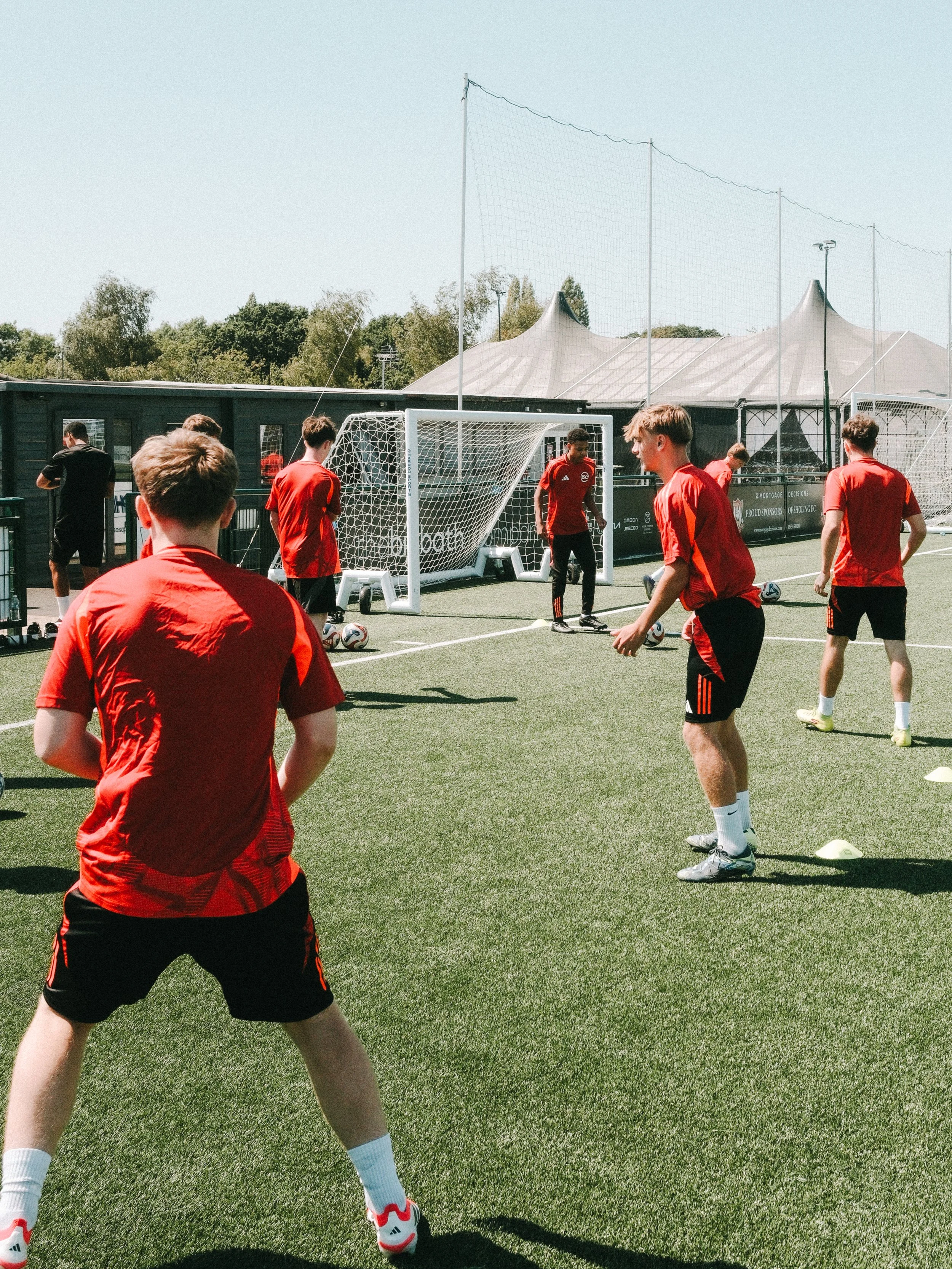 Young soccer players in red jerseys practicing on a synthetic turf field during daytime, with small soccer goals and a large tent structure in the background.