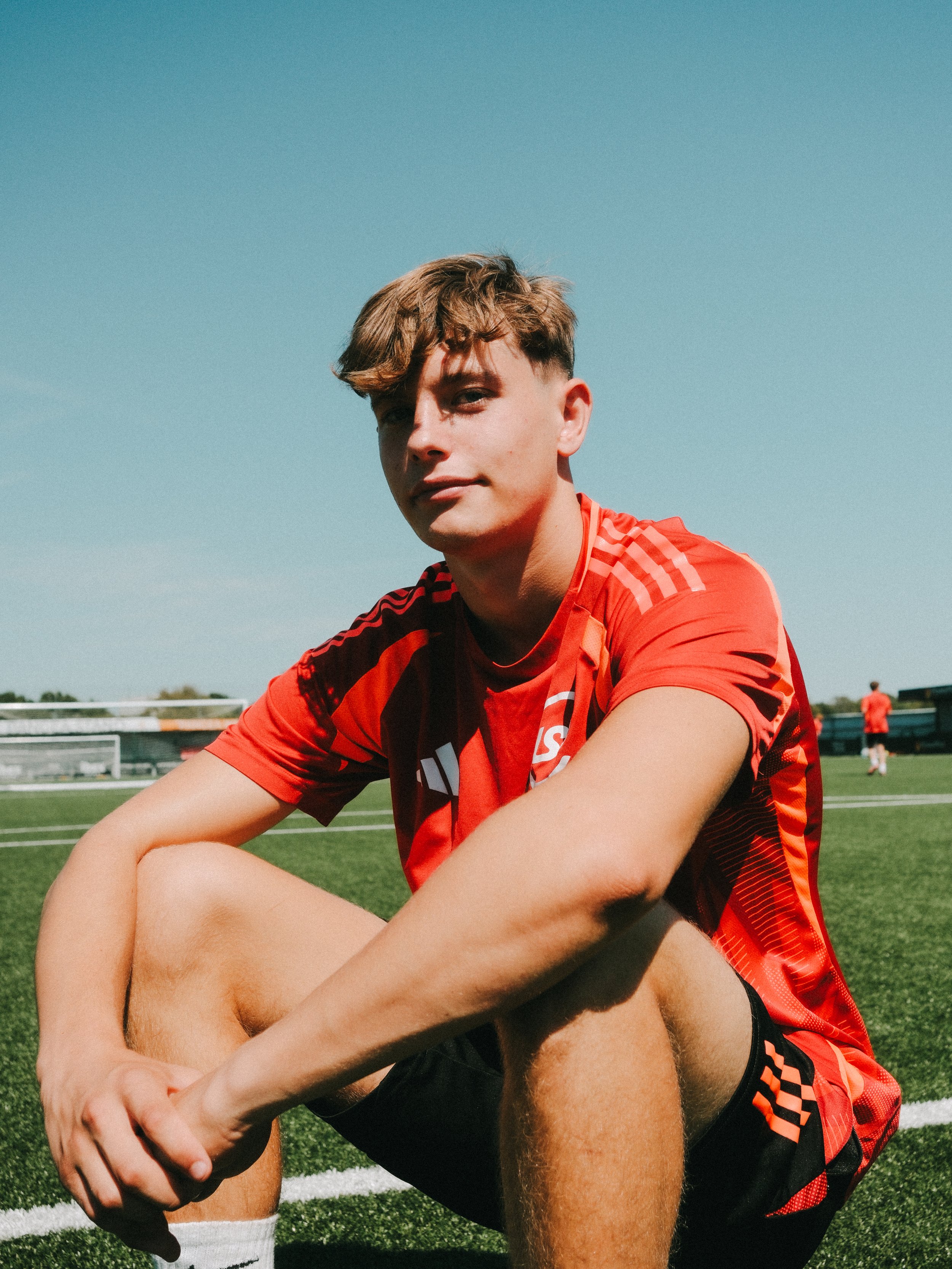 A young male soccer player sitting on a soccer field, wearing a red and black sports uniform, with a blue sky in the background.