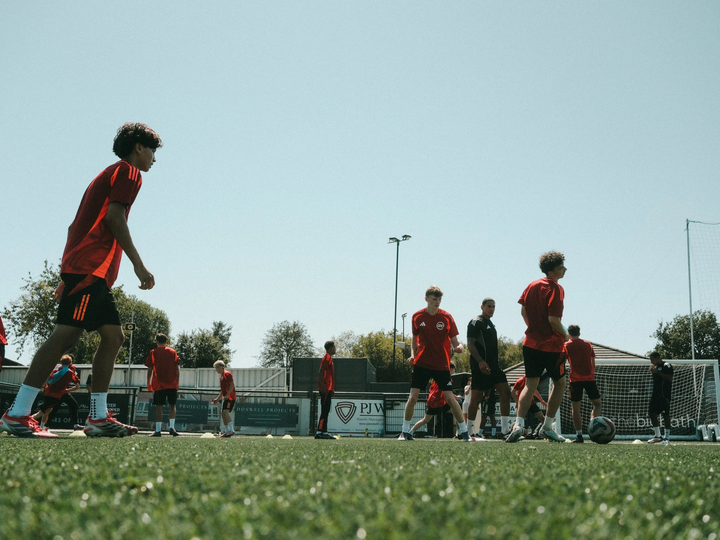 A group of young men in red sports uniforms on a soccer field during practice or a game, with some standing and others walking, under a clear blue sky.