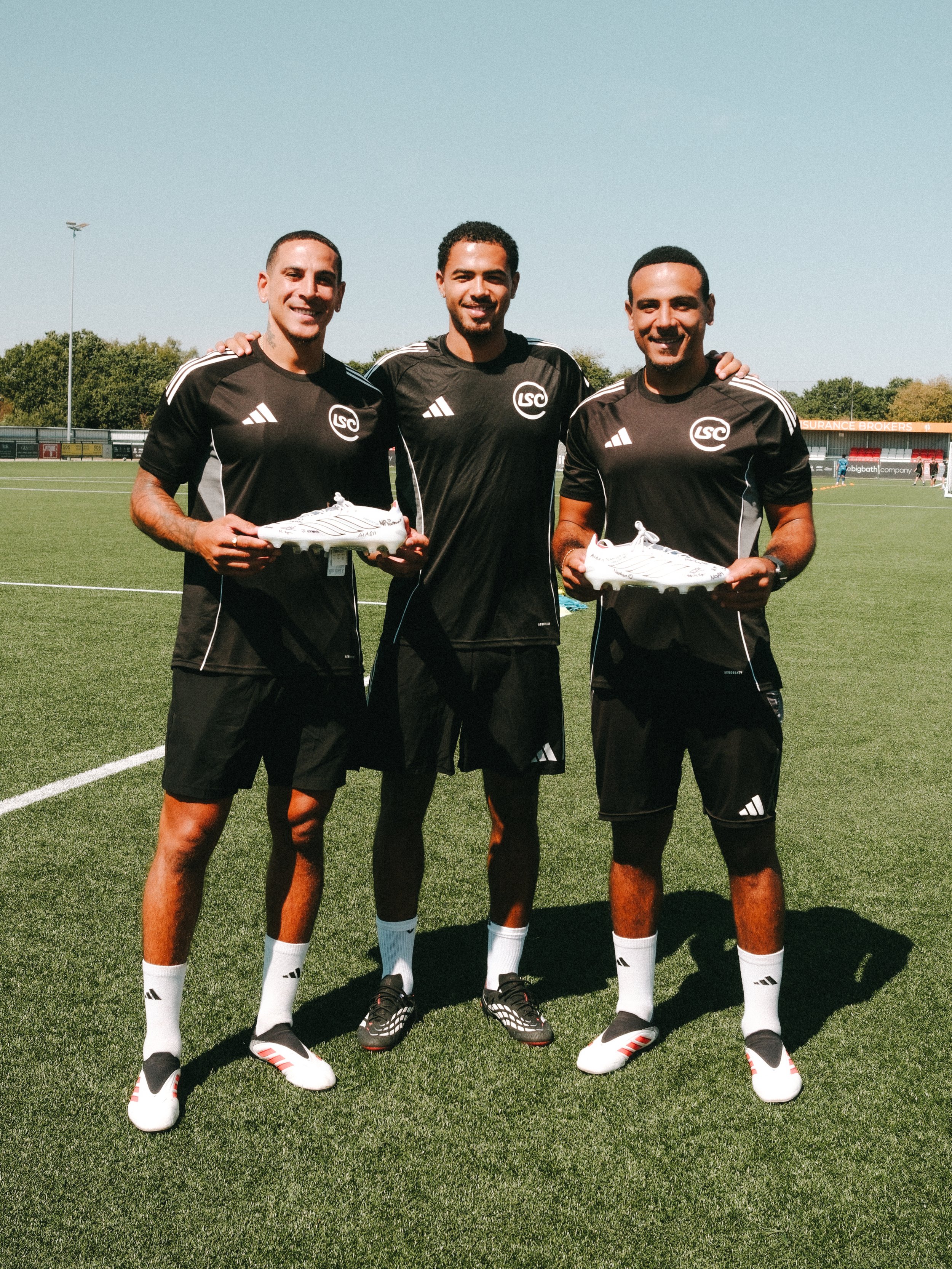 Three football players in black jerseys with white stripes and logos, holding white shoes, standing on a soccer field.