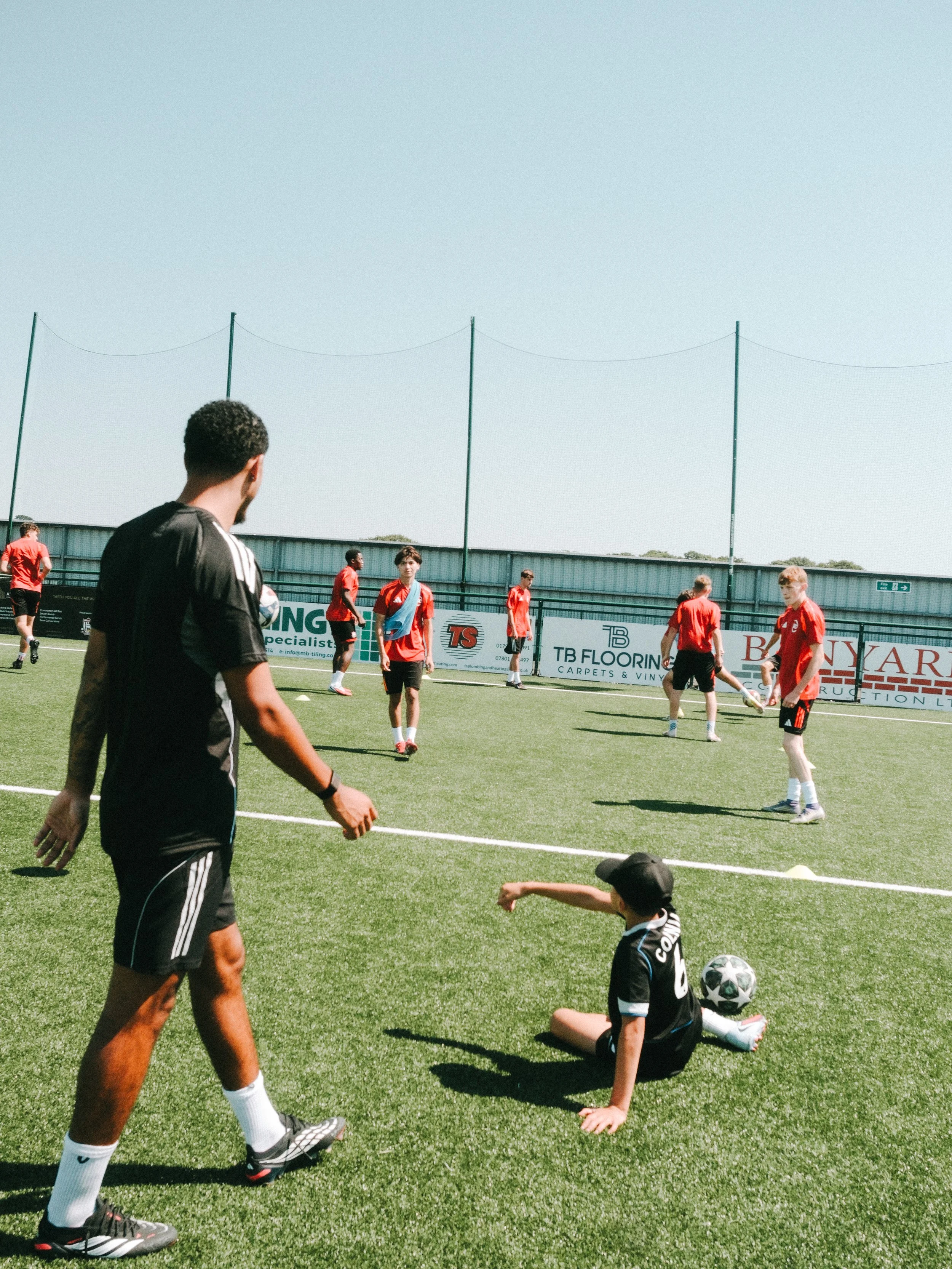 Soccer players practicing on a field with a coach supervising. Some players are standing, while a child in jersey and cap is sitting on the ground with a soccer ball.
