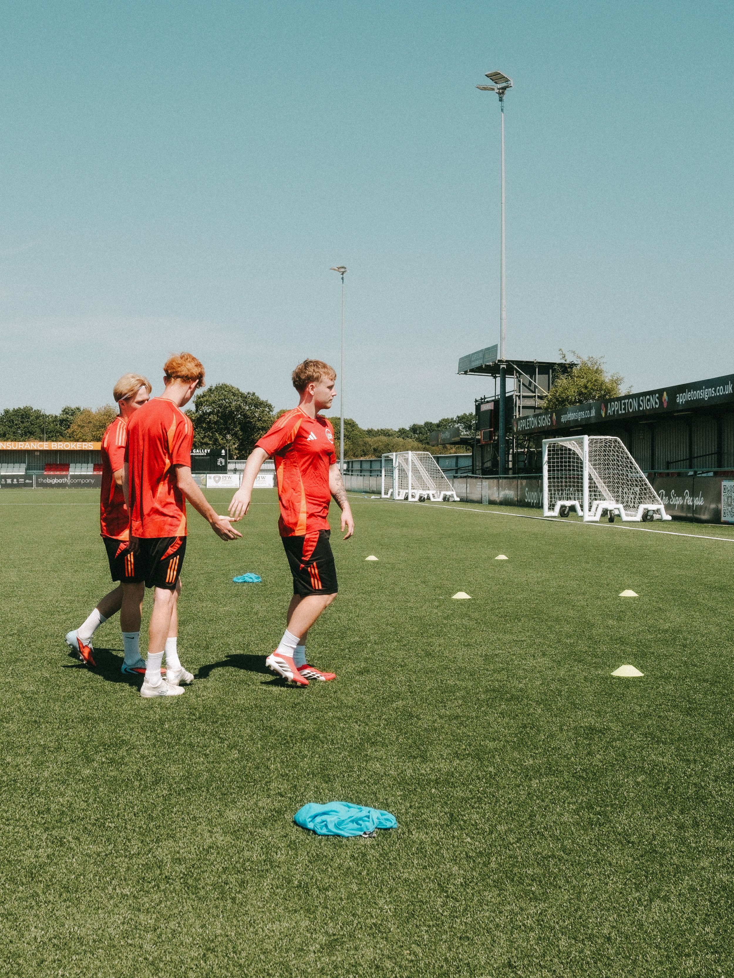 Three young men dressed in red soccer uniforms on a soccer field, practicing drills with small white cones, under a clear blue sky.