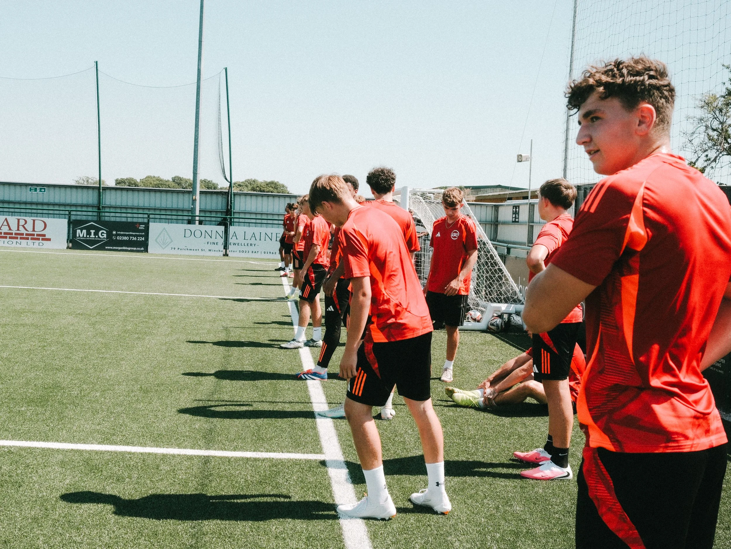 A group of young male soccer players in red uniforms stand in a line on the sidelines of a soccer field, preparing for the game, with a green fence and advertisements in the background on a sunny day.