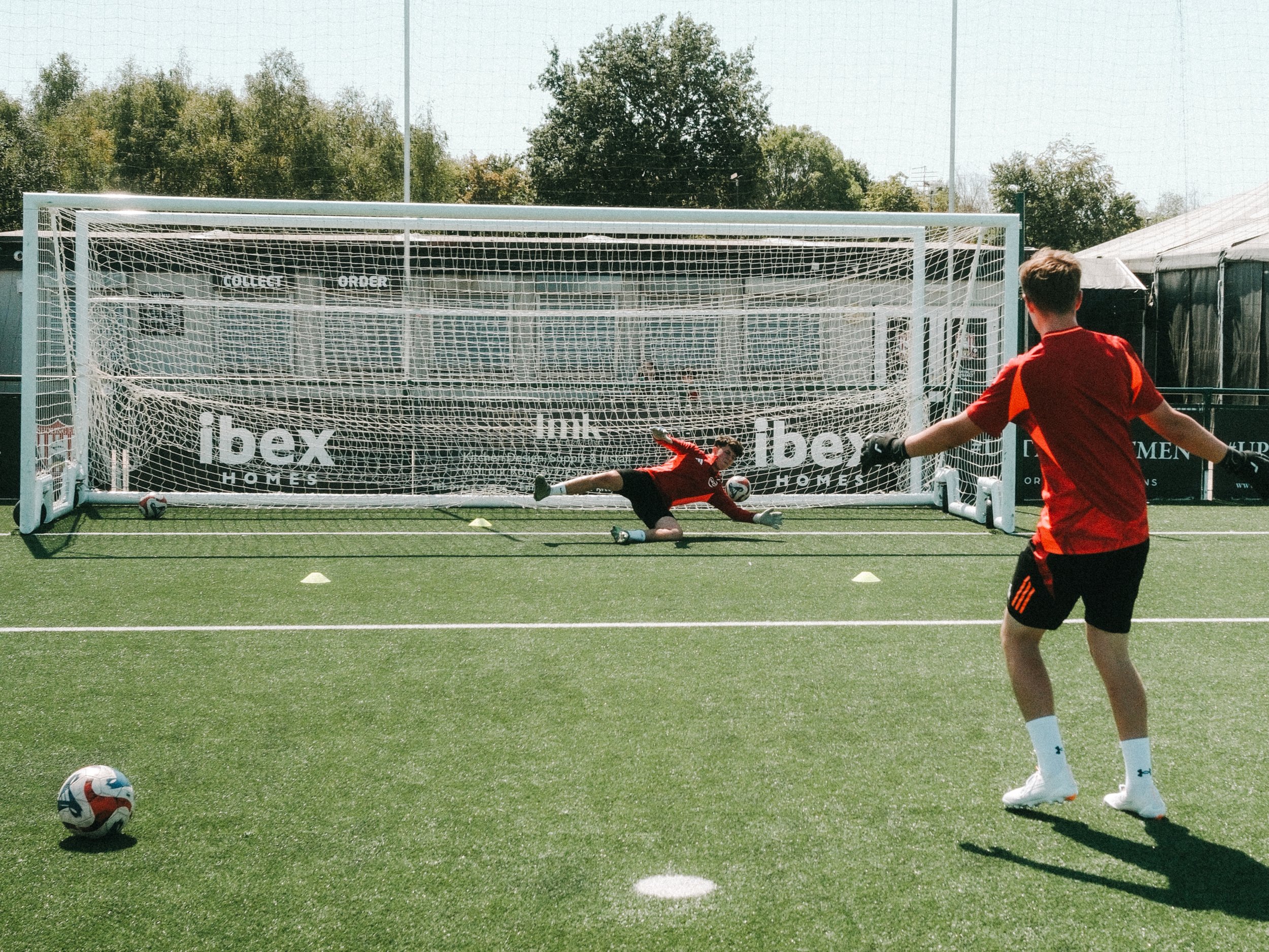 A soccer player in a red uniform attempting to score a goal while a goalkeeper in a black uniform dives to block the shot in a soccer goal with white posts and netting on a green field.