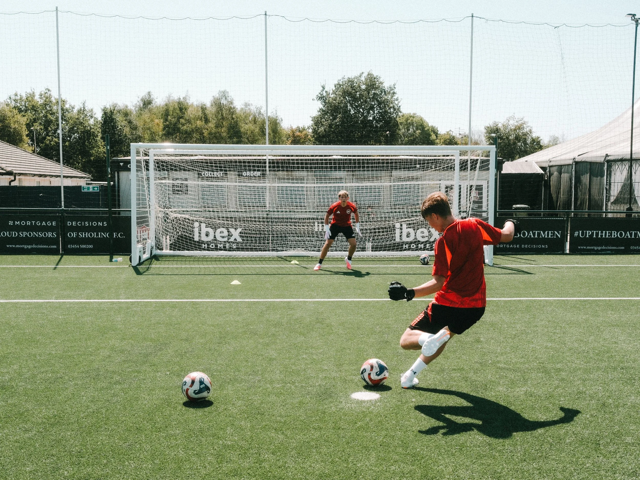 A soccer practice session with players kicking balls on a field. A goalkeeper stands in front of the goal, and a player in red is kicking a soccer ball. Additional soccer balls are on the ground, and the goal has advertising banners on it.