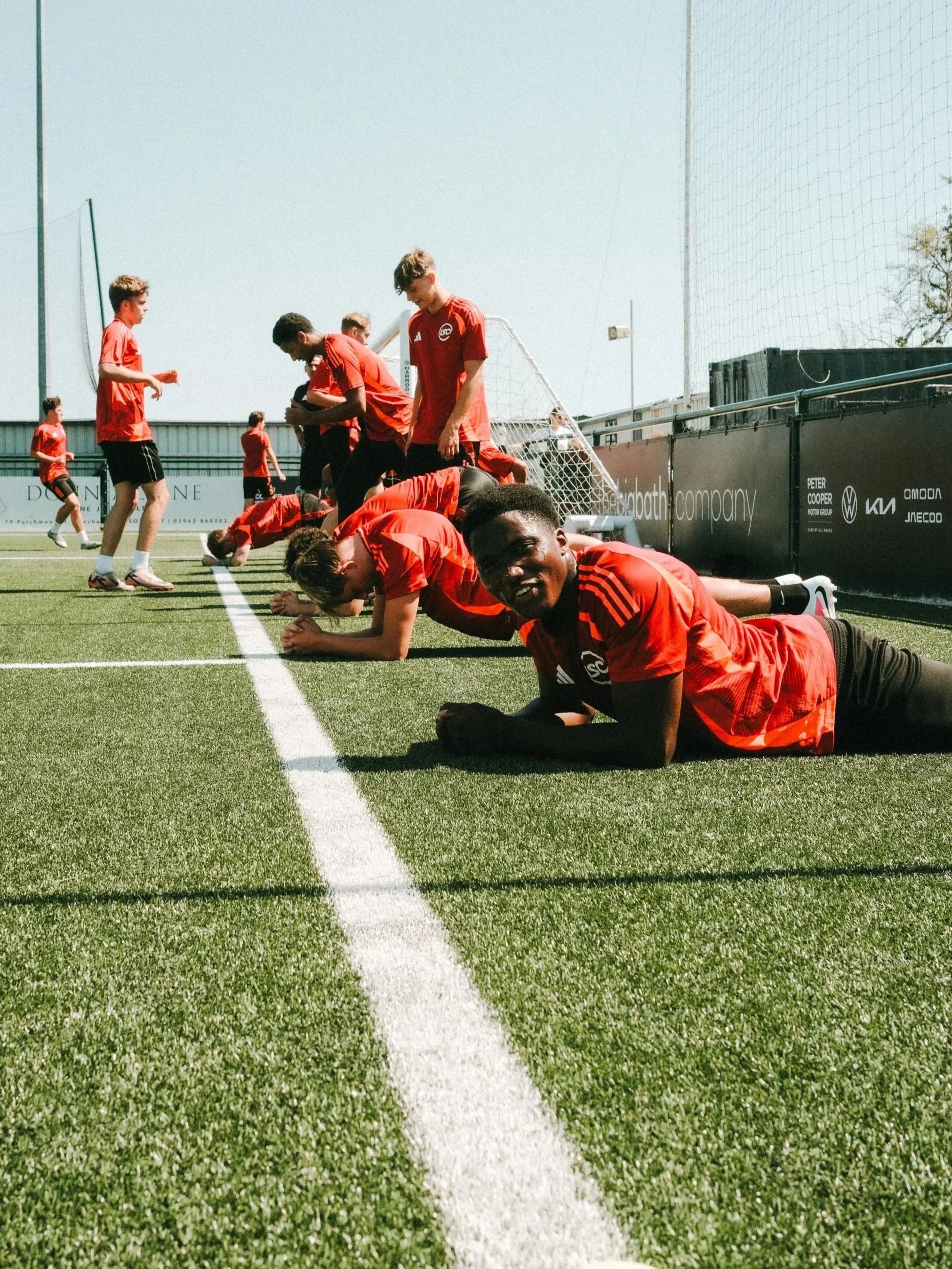 Soccer players in red jerseys practicing on a field, with some standing and others on the ground, near a goalpost.