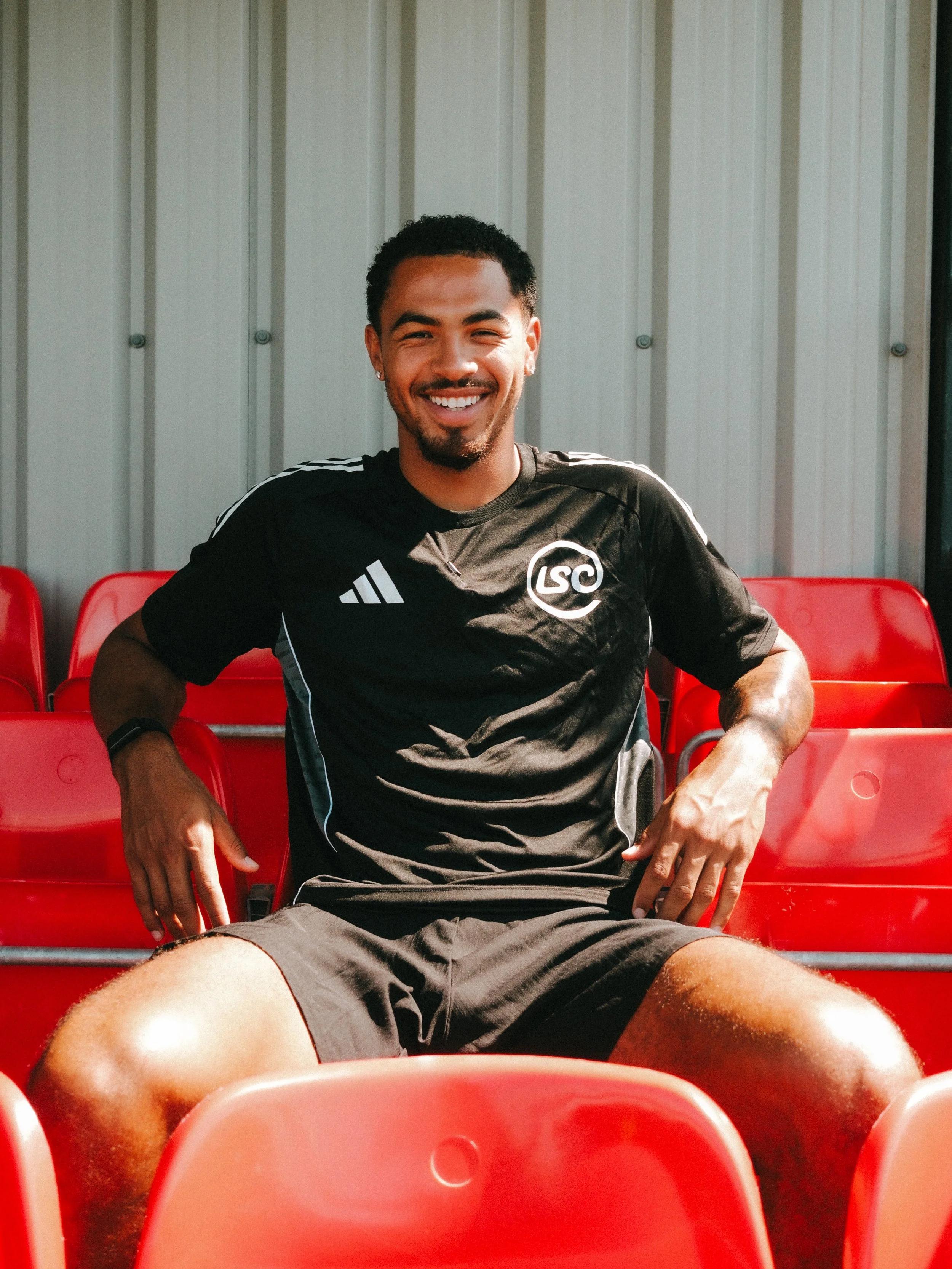 Young man sitting on red stadium seats, smiling at camera, wearing a black athletic shirt with 'LSC' logo, shorts, and earrings.