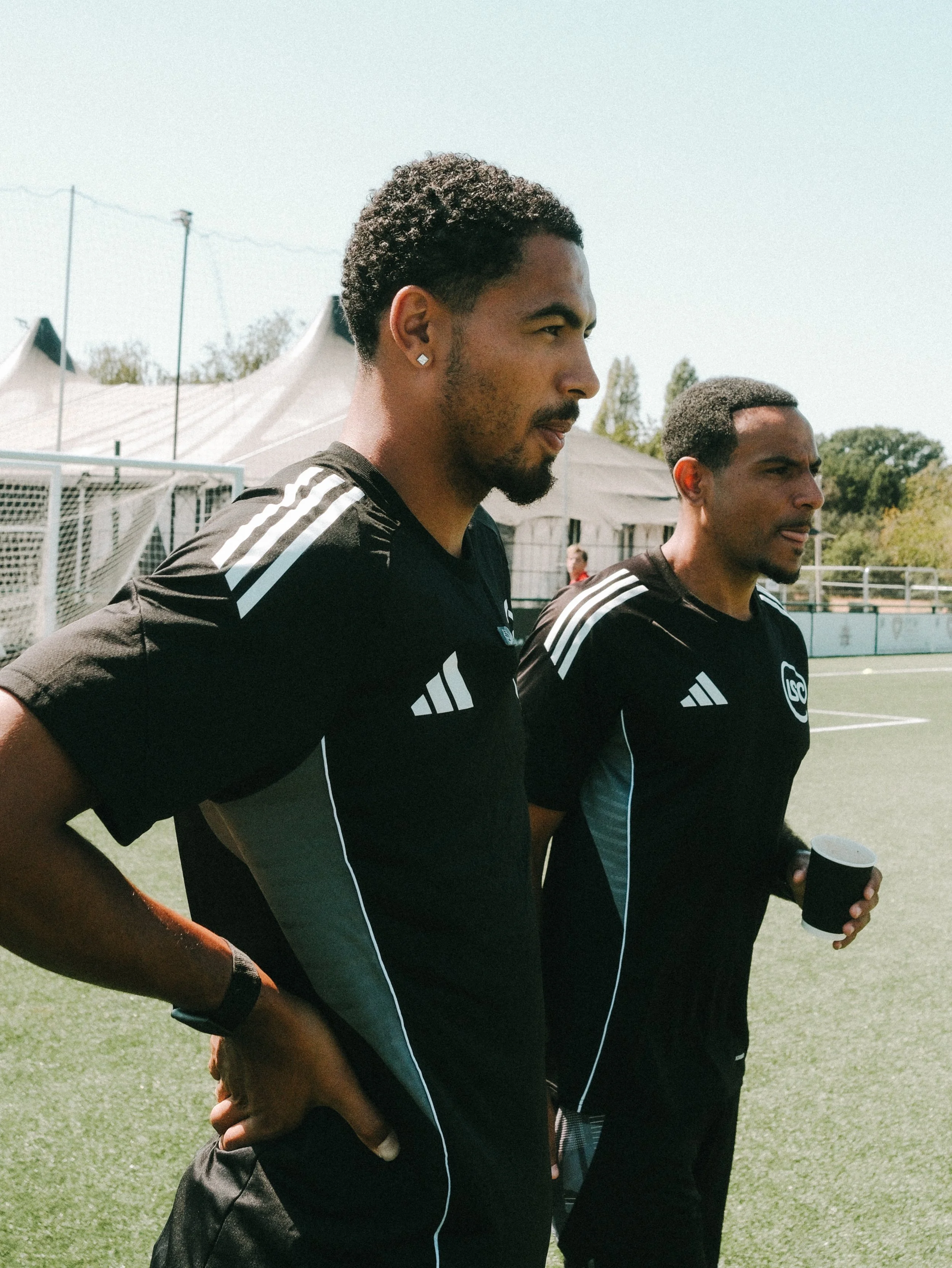 Two male soccer players standing side by side on a field, wearing black jerseys with white stripes on the shoulders, one holding a black coffee cup, with a tent and trees in the background on a sunny day.