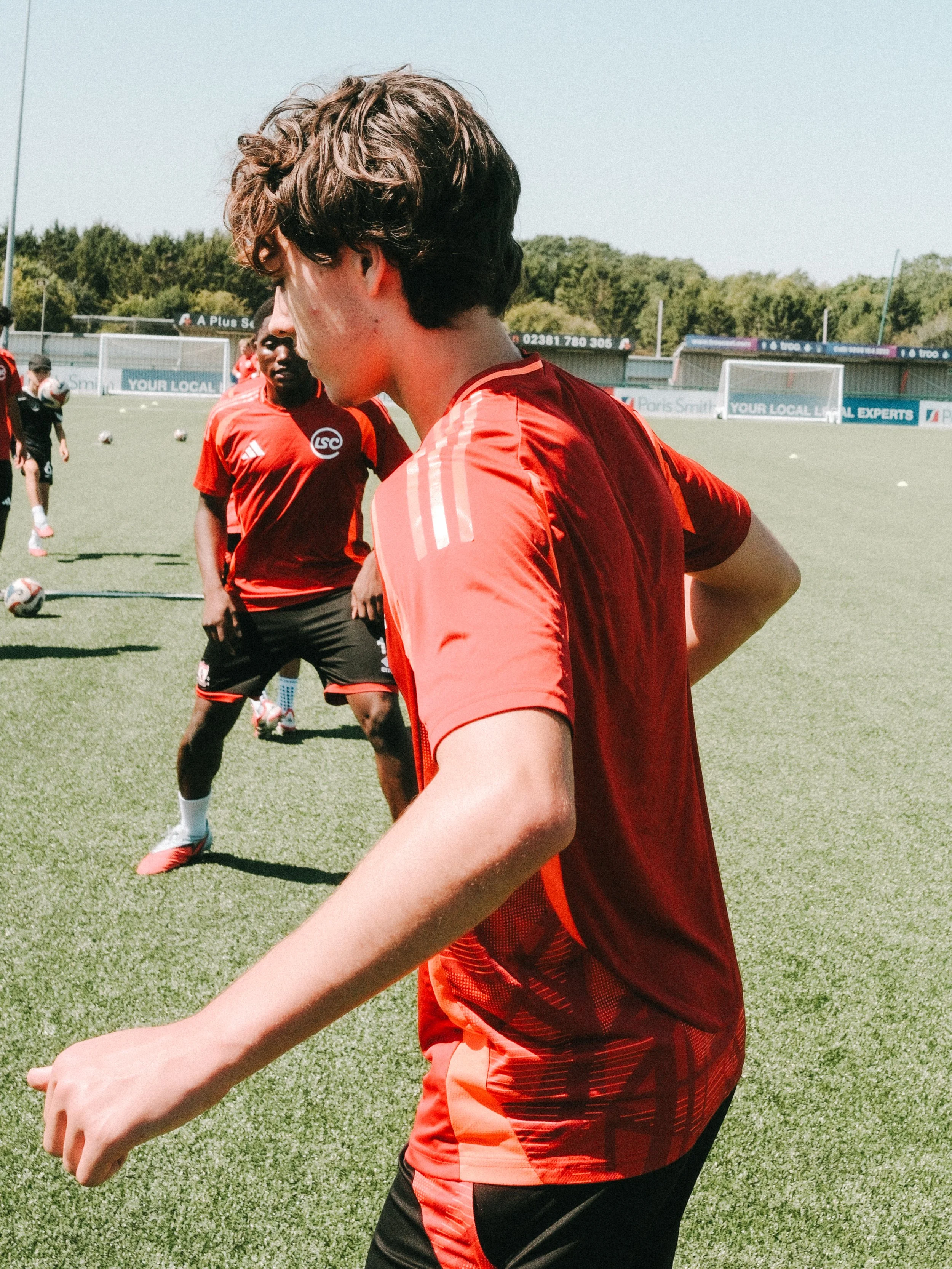 A group of young men wearing red sports jerseys and black shorts on a soccer field during daytime, with soccer balls on the ground, practicing or preparing for a game.
