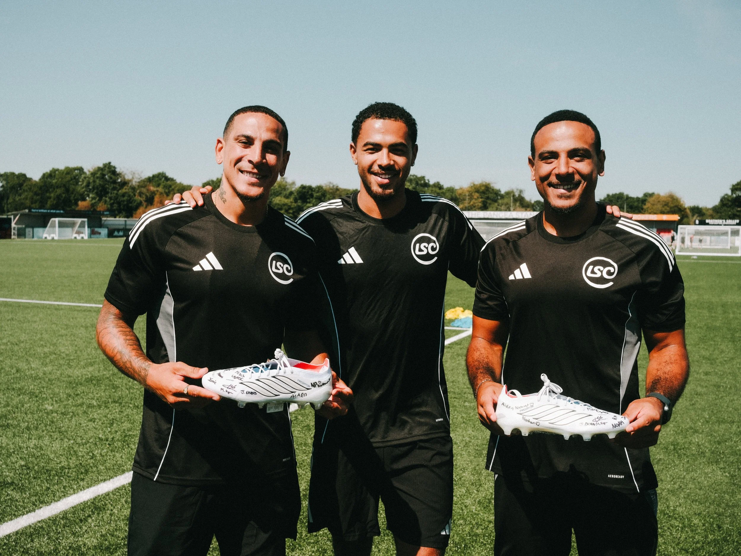 Three smiling soccer players in black jerseys standing on a soccer field, holding signed white soccer cleats.