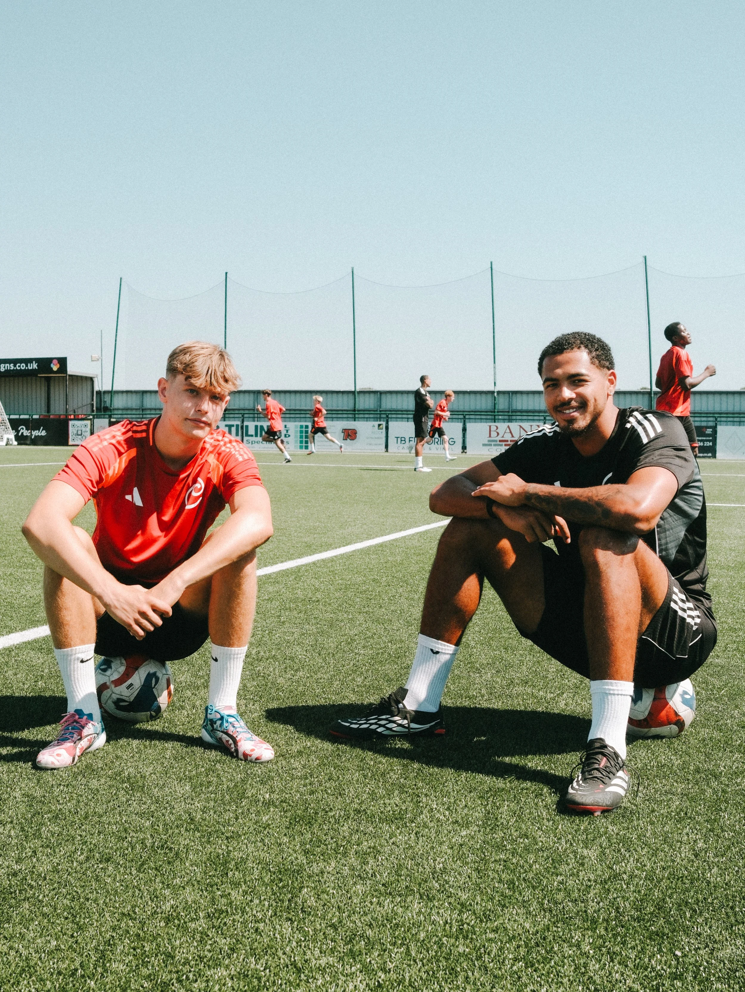 Two soccer players crouching on a field, with teammates warming up in the background, under a clear blue sky.