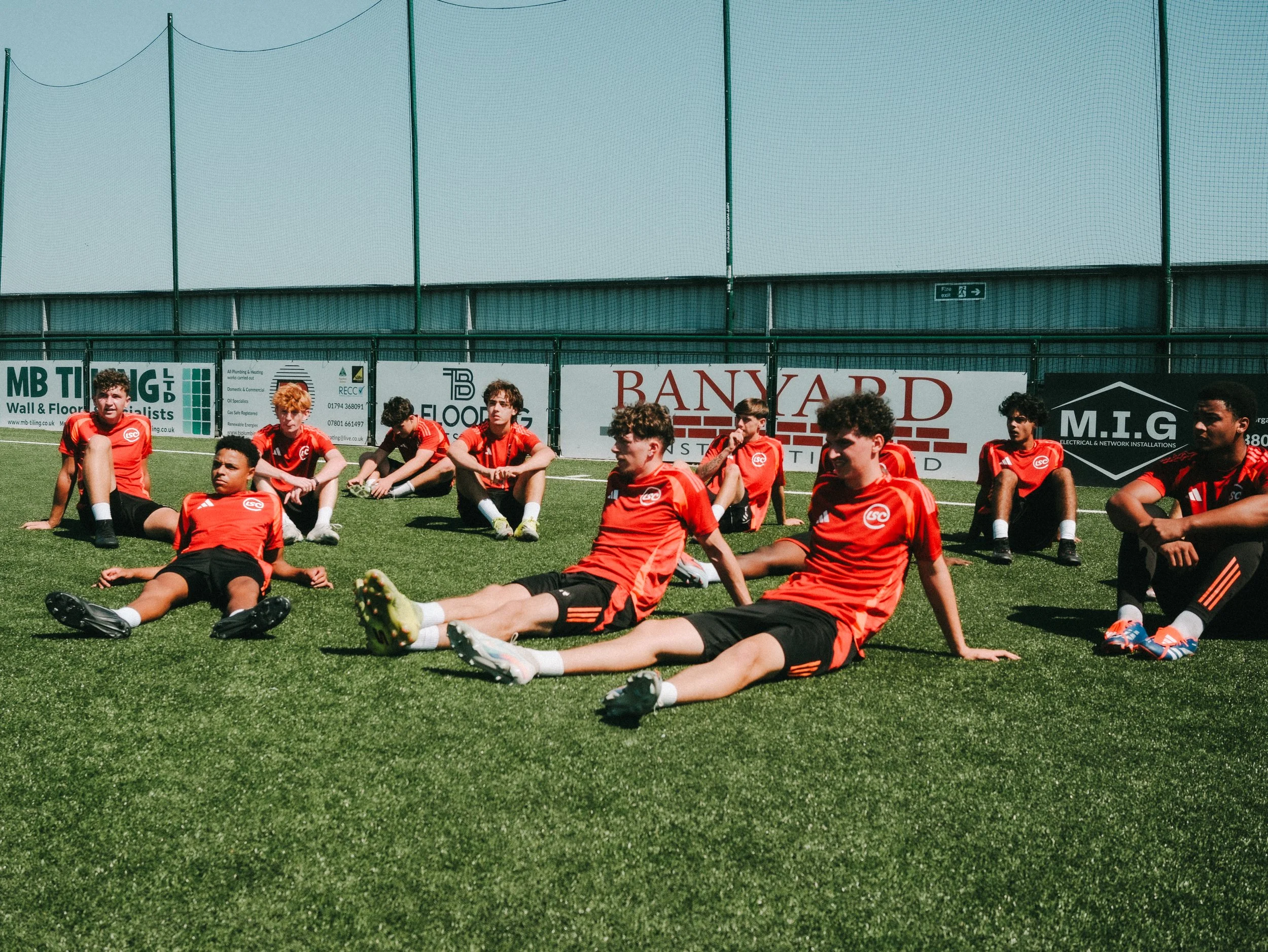 A group of young soccer players sitting on a green field, wearing red uniforms with black shorts, with advertising banners behind them and a pale blue sky overhead.