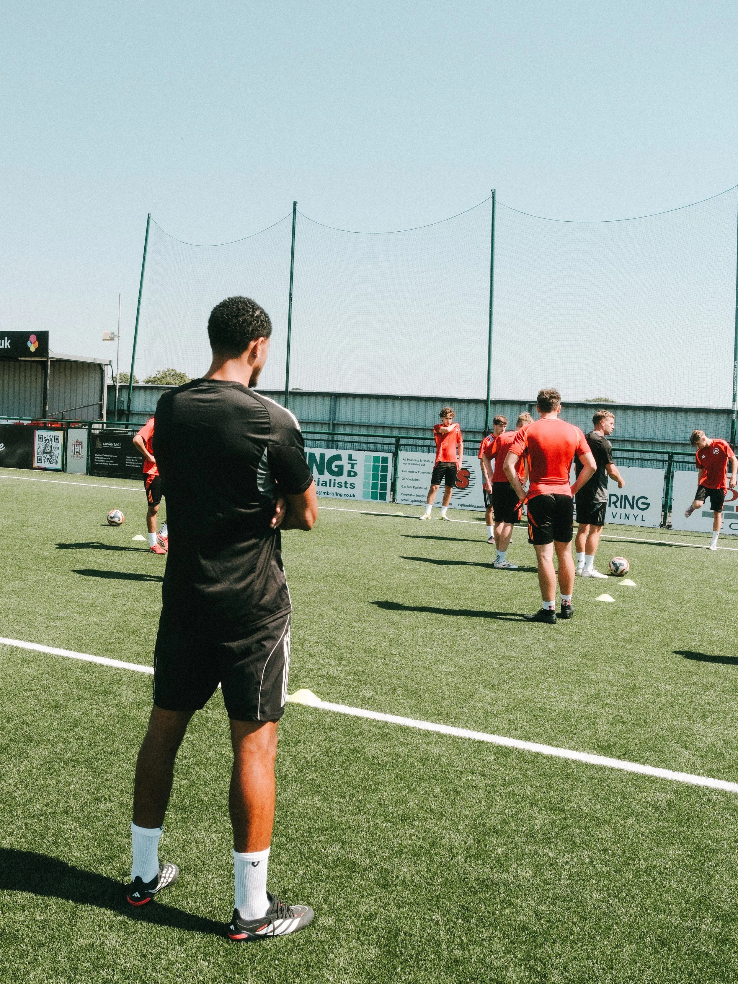 A soccer coach or trainer standing on the field with arms crossed, observing a group of players in red and black uniforms during practice on a sunny day.