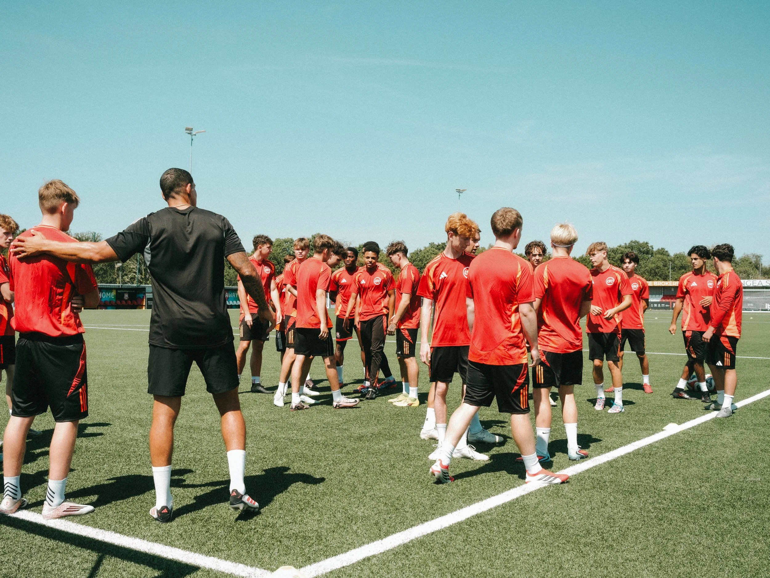 Group of young soccer players in red uniforms gathered on the field during a practice or game, with a coach speaking to them under a clear blue sky.