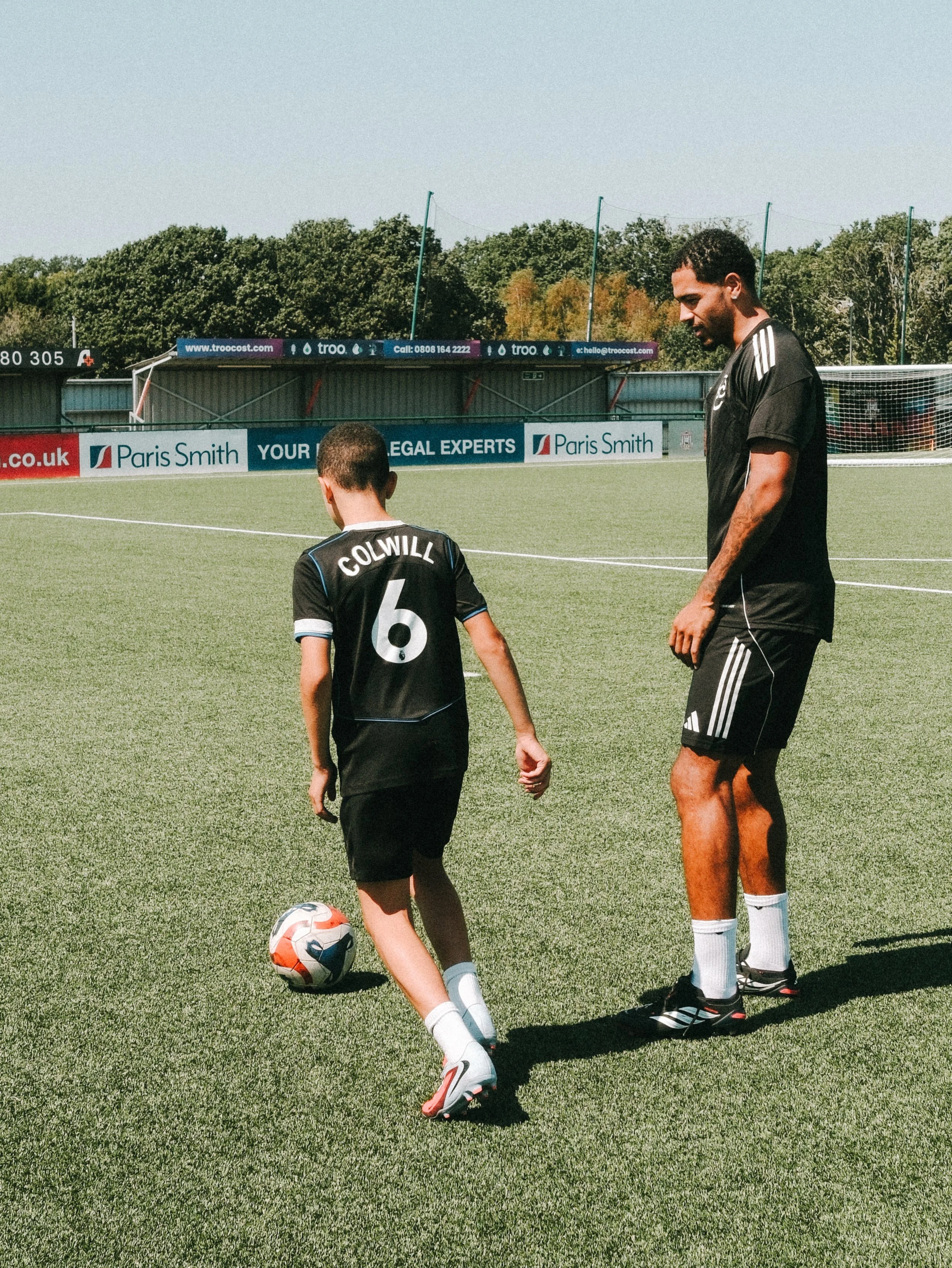 A young boy with the name 'Colwill' on his football jersey practicing soccer on a field with an adult male, who is wearing a black sports shirt and shorts. The boy is about to kick a soccer ball, and they are both standing on the grass with a goalpos