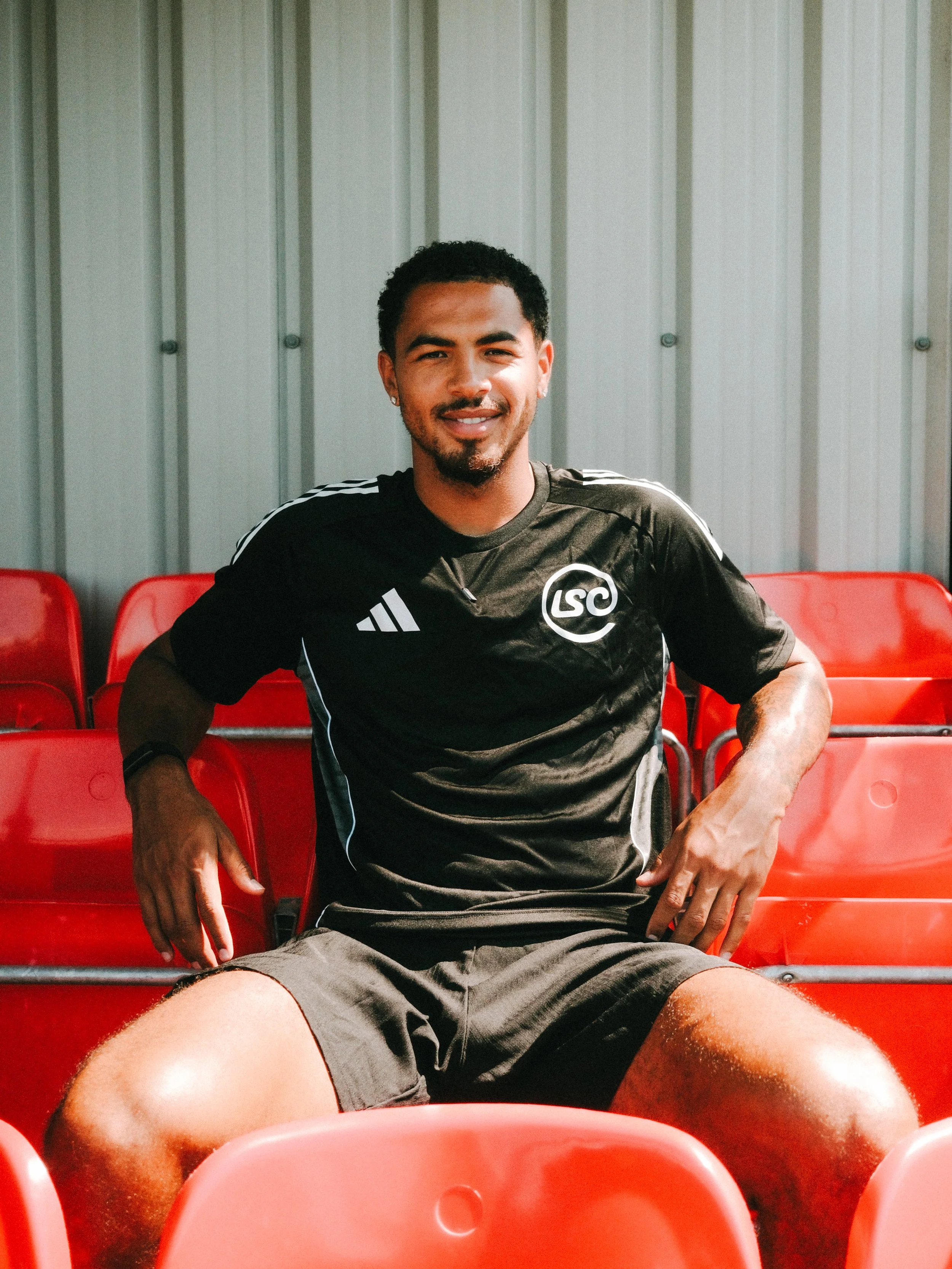 A man sitting in a sports stadium on red seats, wearing a black athletic jersey with the logo 'LSC' and smiling.