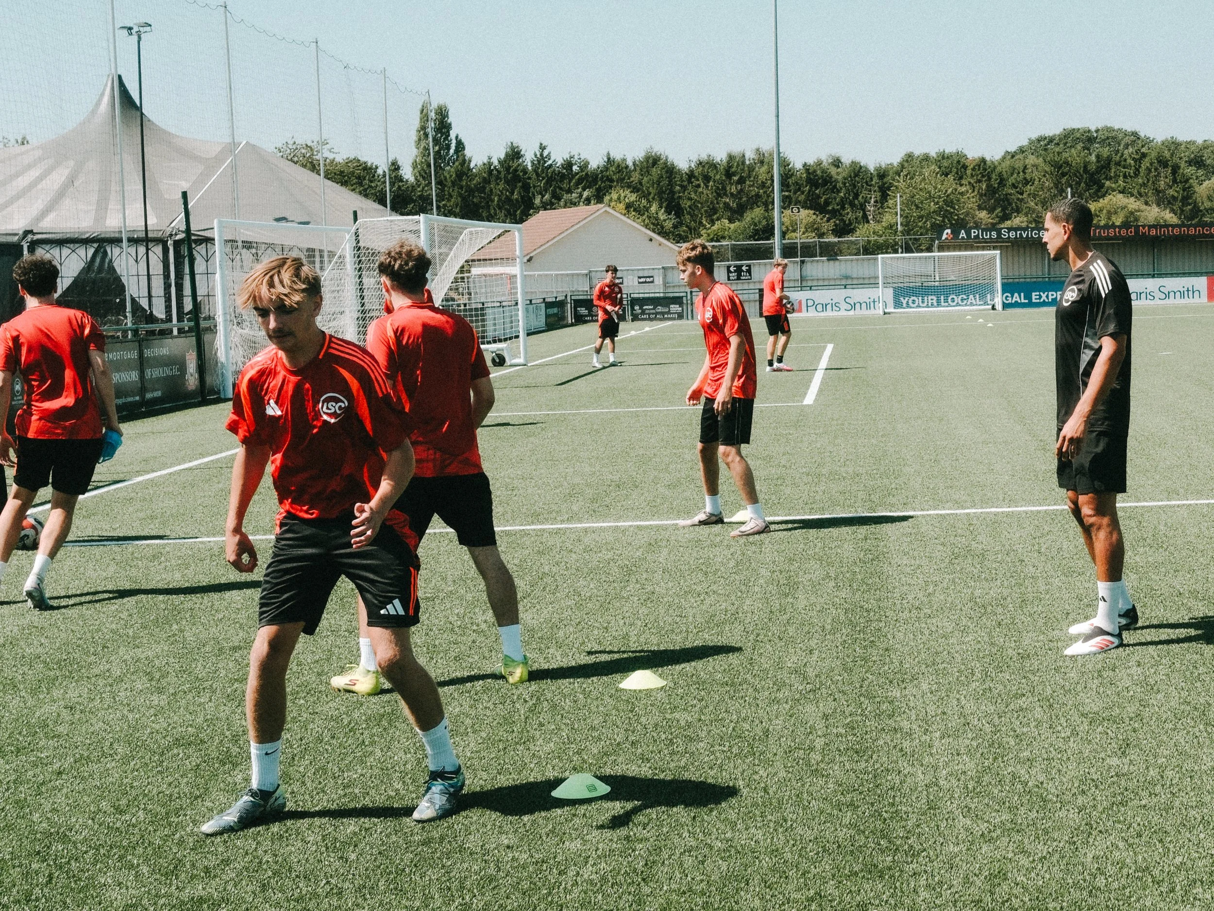 A group of young soccer players practicing on a field with cones, wearing red and black jerseys, and a coach or referee wearing a black shirt.