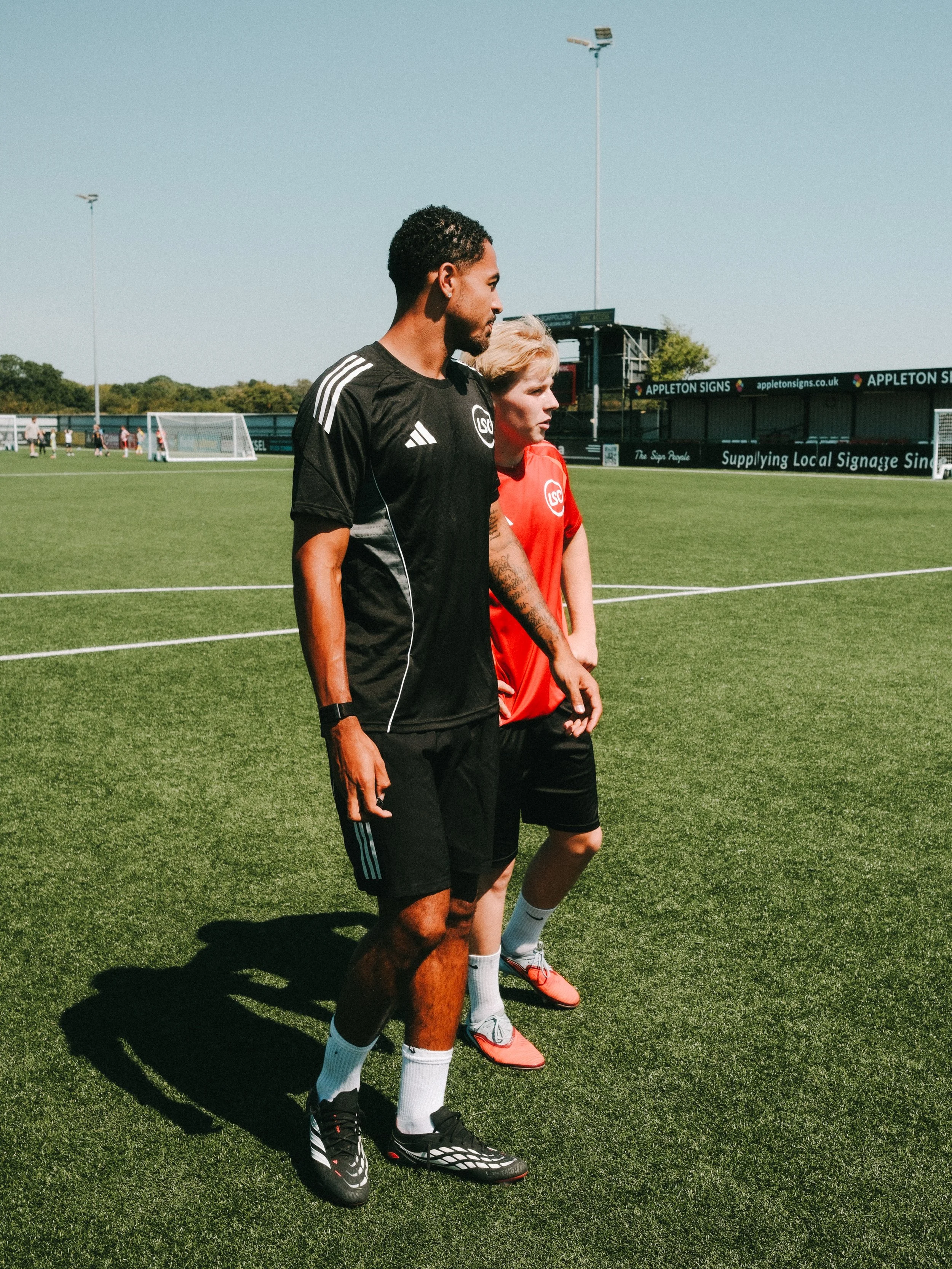 Two soccer players walking on a soccer field, one wearing a black jersey and the other a red jersey. The field is green with goalposts in the background and a sign advertising local signage.
