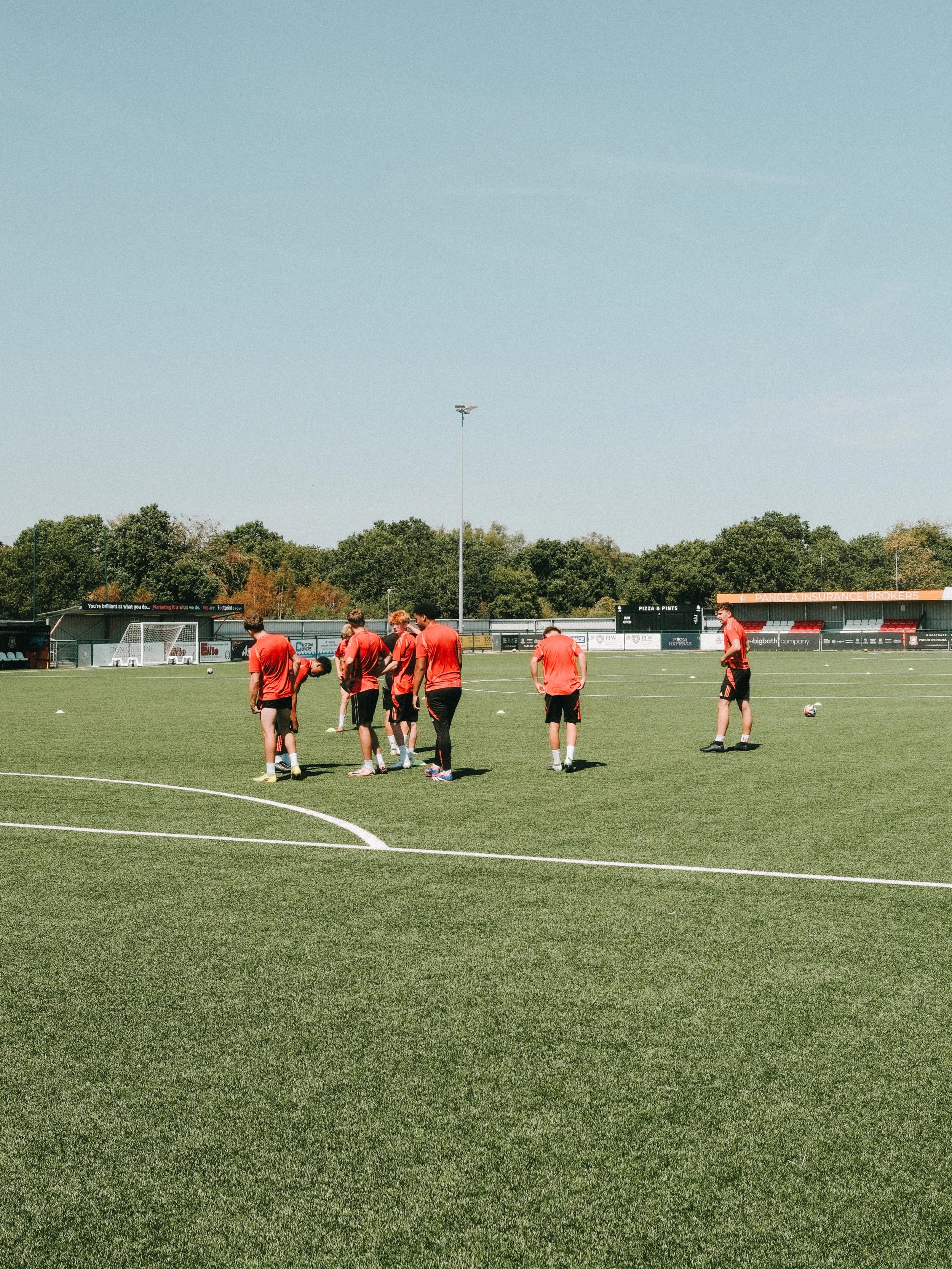 A group of soccer players in red uniforms on a soccer field during practice or a game, with trees in the background and a scoreboard visible.