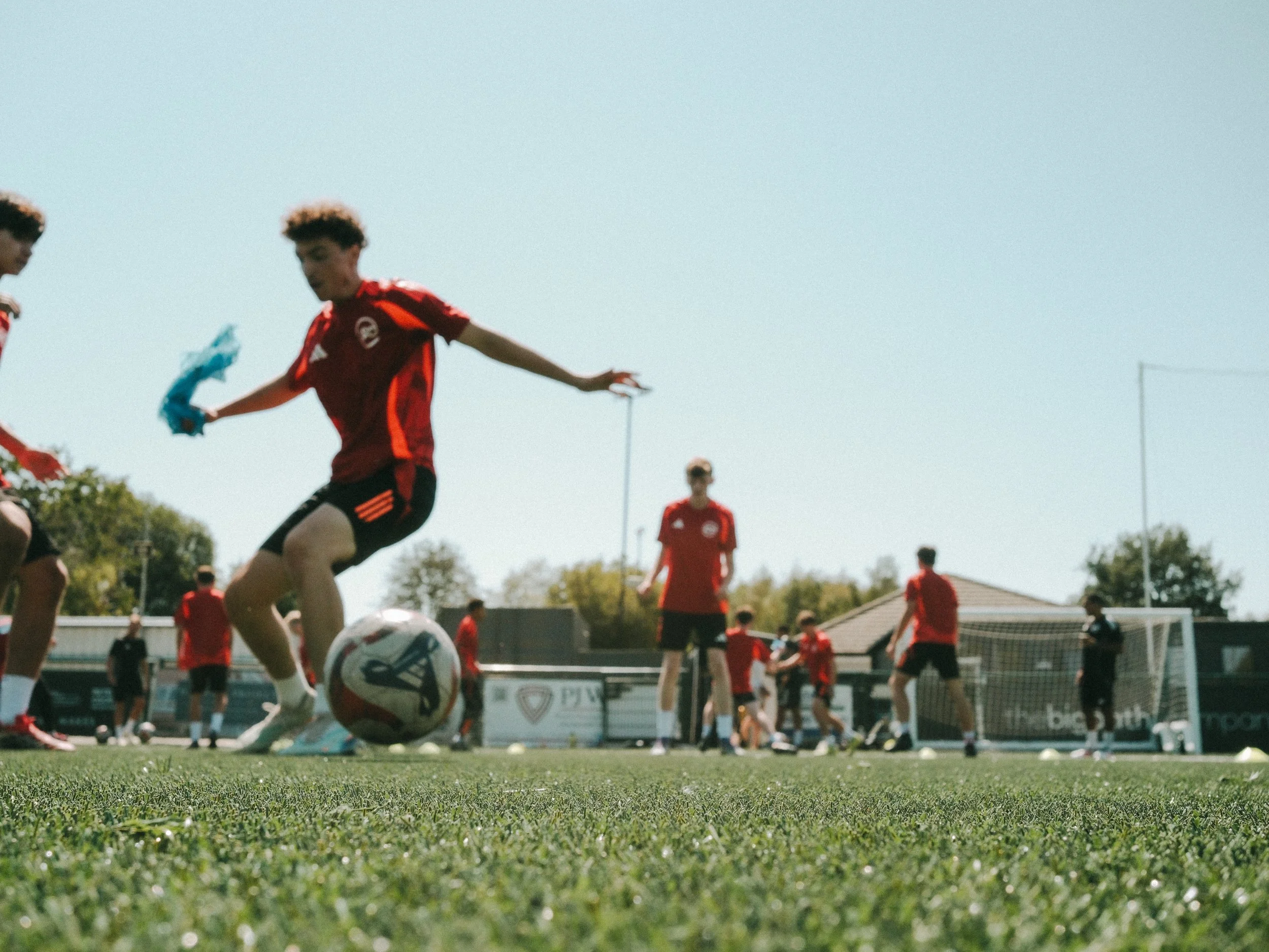 Youth soccer players in red uniforms playing a game on a green field, with one player kicking a soccer ball and others watching, under a clear blue sky.