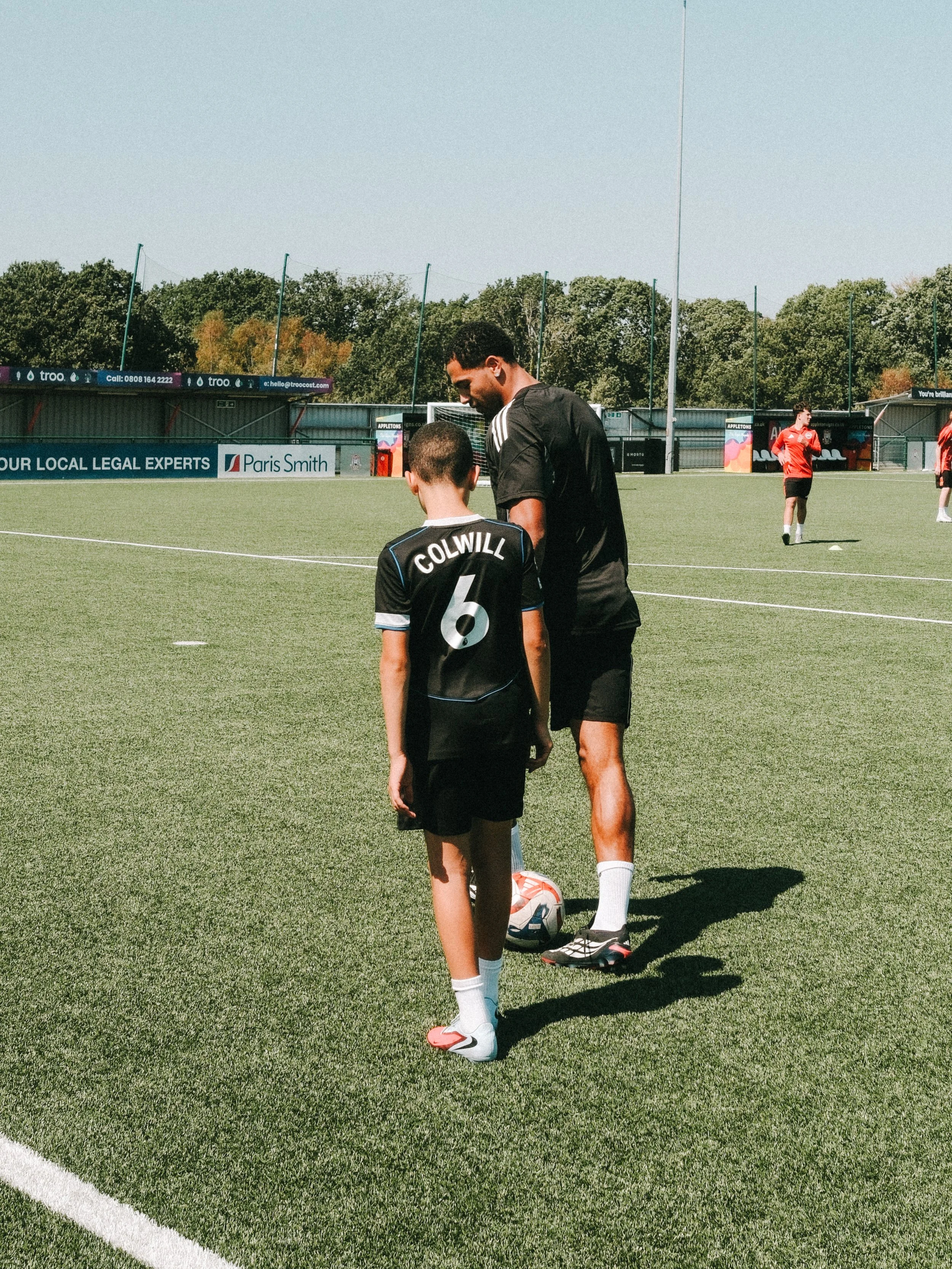 A soccer coach giving instructions to a young player during practice on a green outdoor soccer field with other players and trees in the background.