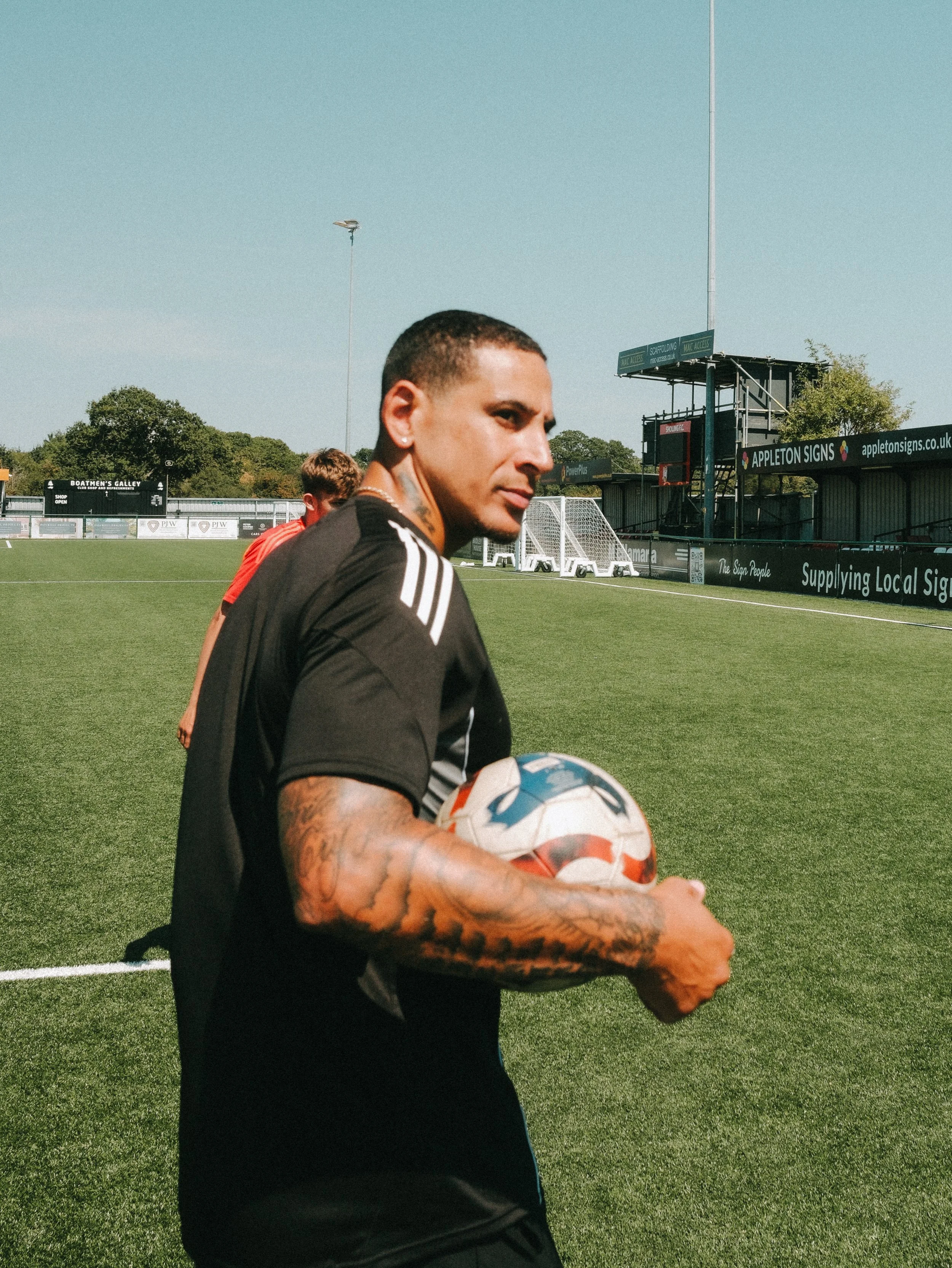 A man with tattoos on his arms holding a soccer ball on a soccer field with a scoreboard and stands in the background.
