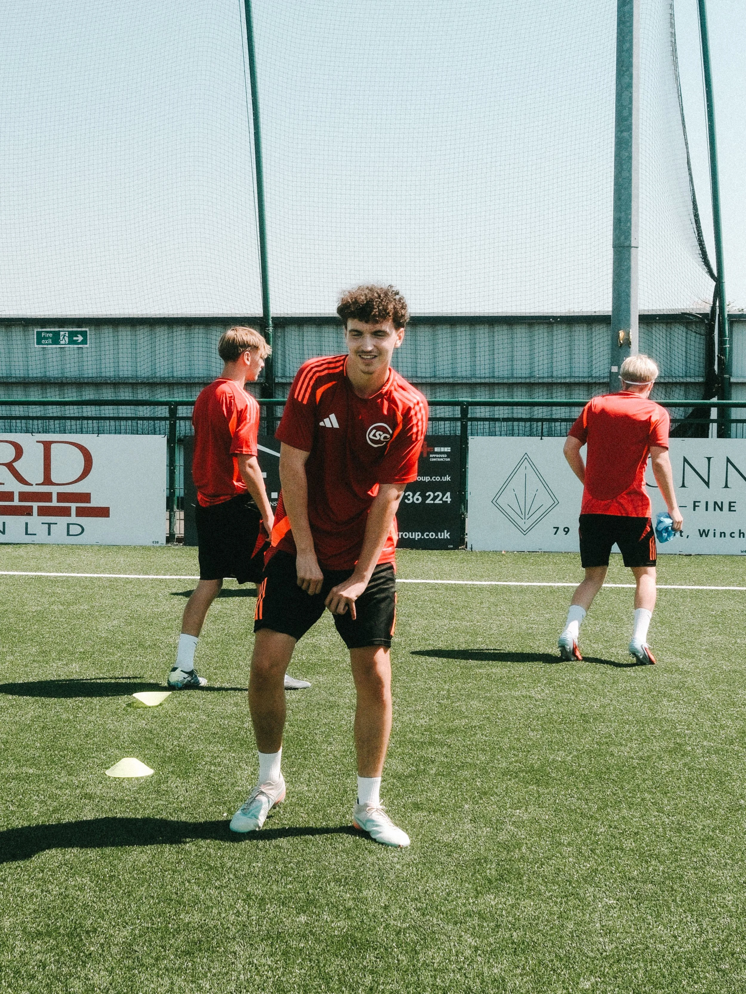 Young male soccer player in a red and black training kit standing on a soccer field, with other players in the background, training cones on the ground, and a fence with advertising banners behind him.