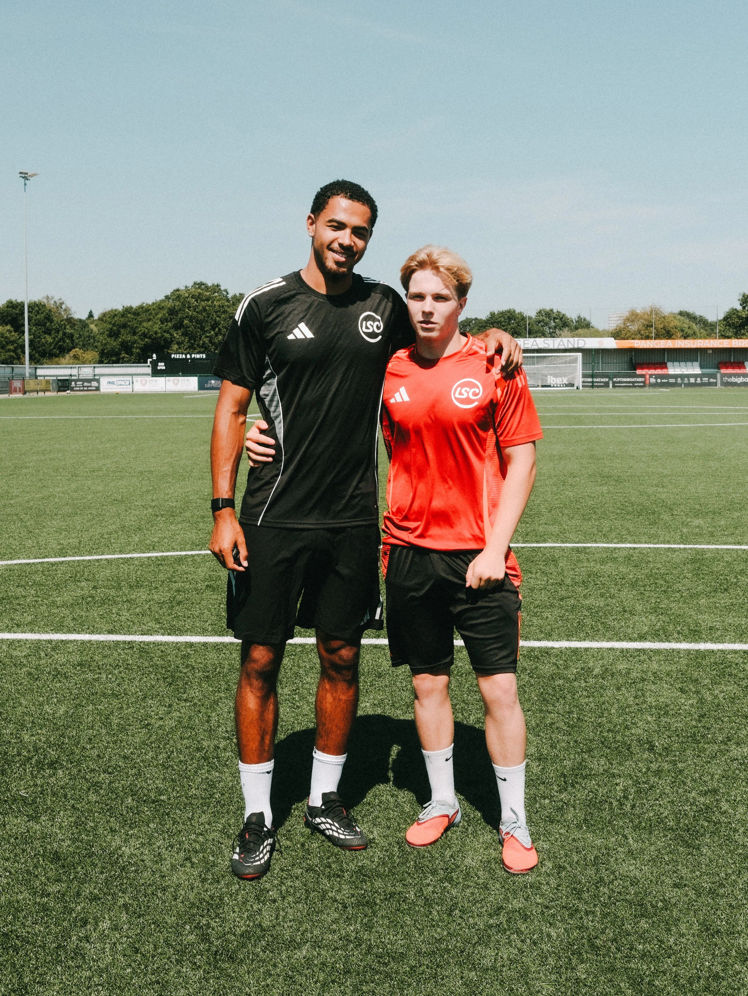 Two soccer players, one in a black jersey and the other in a red jersey, standing on a soccer field with their arms around each other.