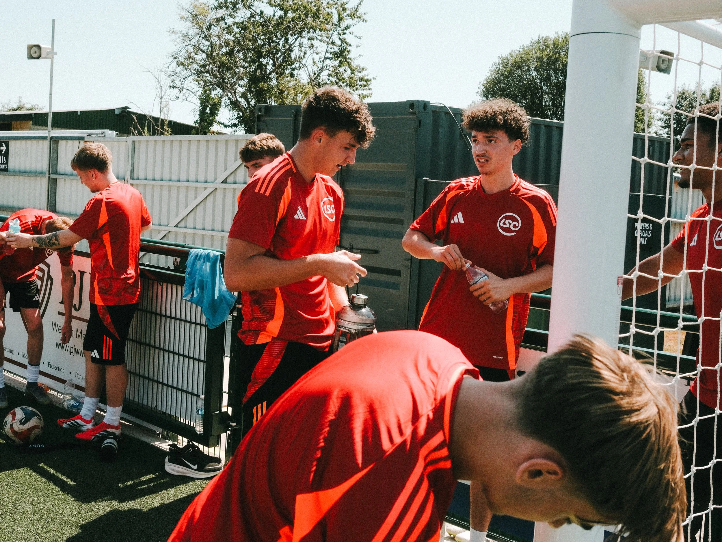 A group of young male soccer players in red jerseys gathered near the sideline of a soccer field, some holding water bottles, during daytime.