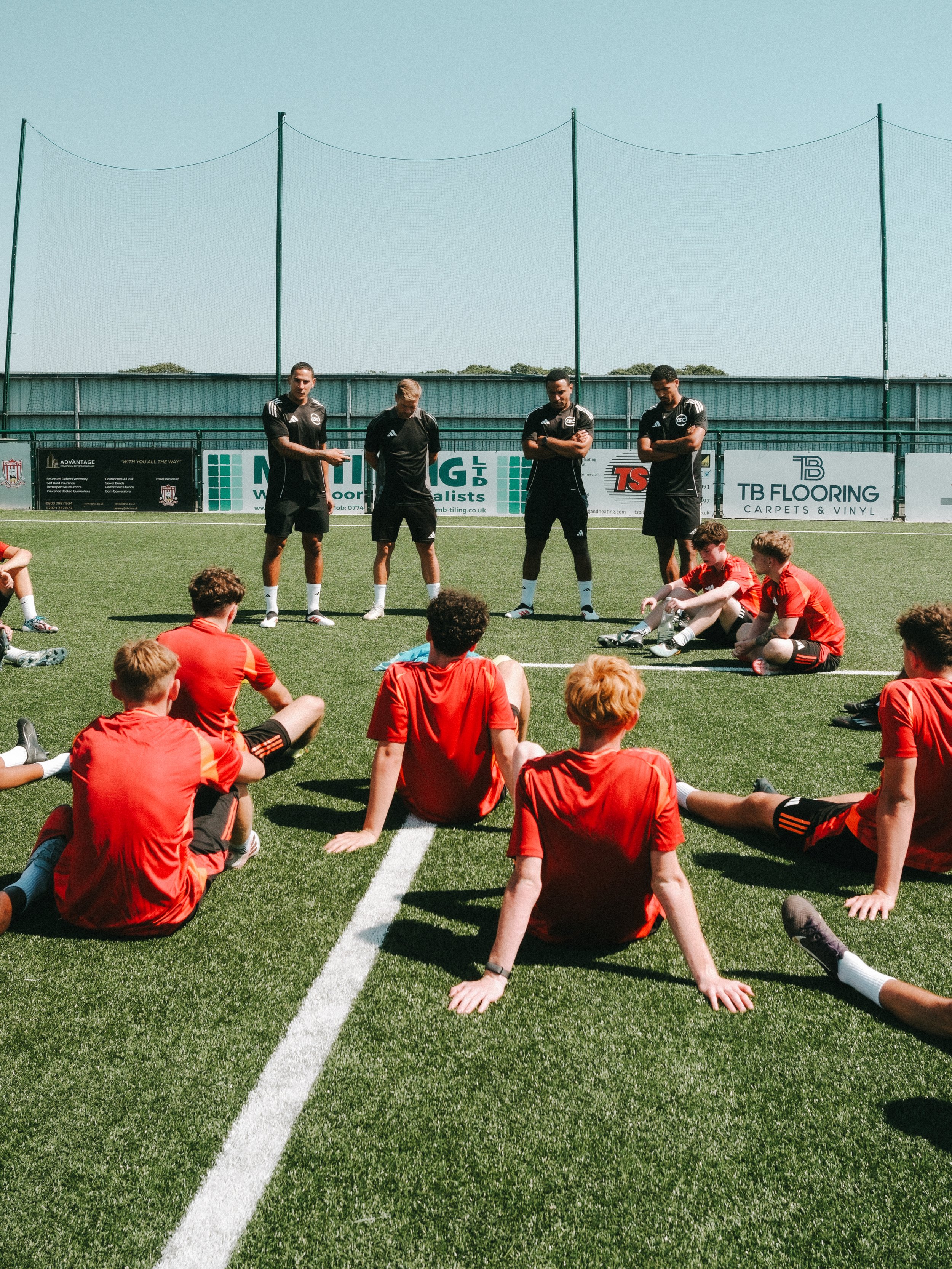 Youth soccer team sitting on the grass field, listening to coach with four players standing in front, outdoors under clear sky.