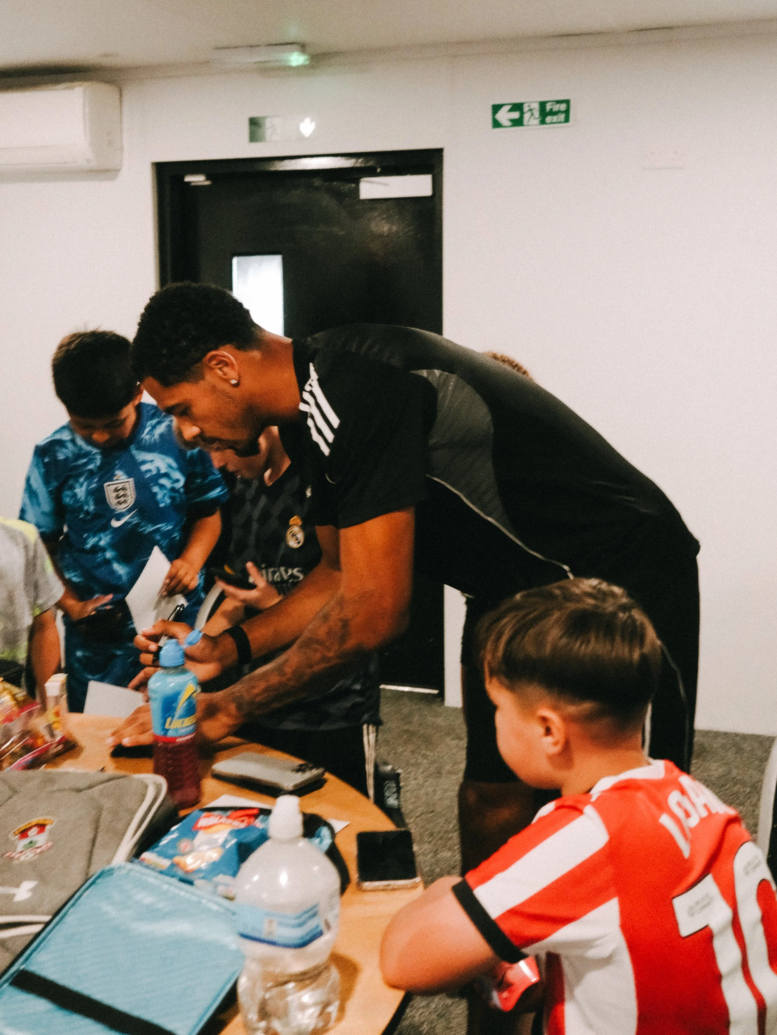People gathered around a table signing autographs, with children also present. The scene appears to be in an indoor setting, possibly a sports event or fan meeting.