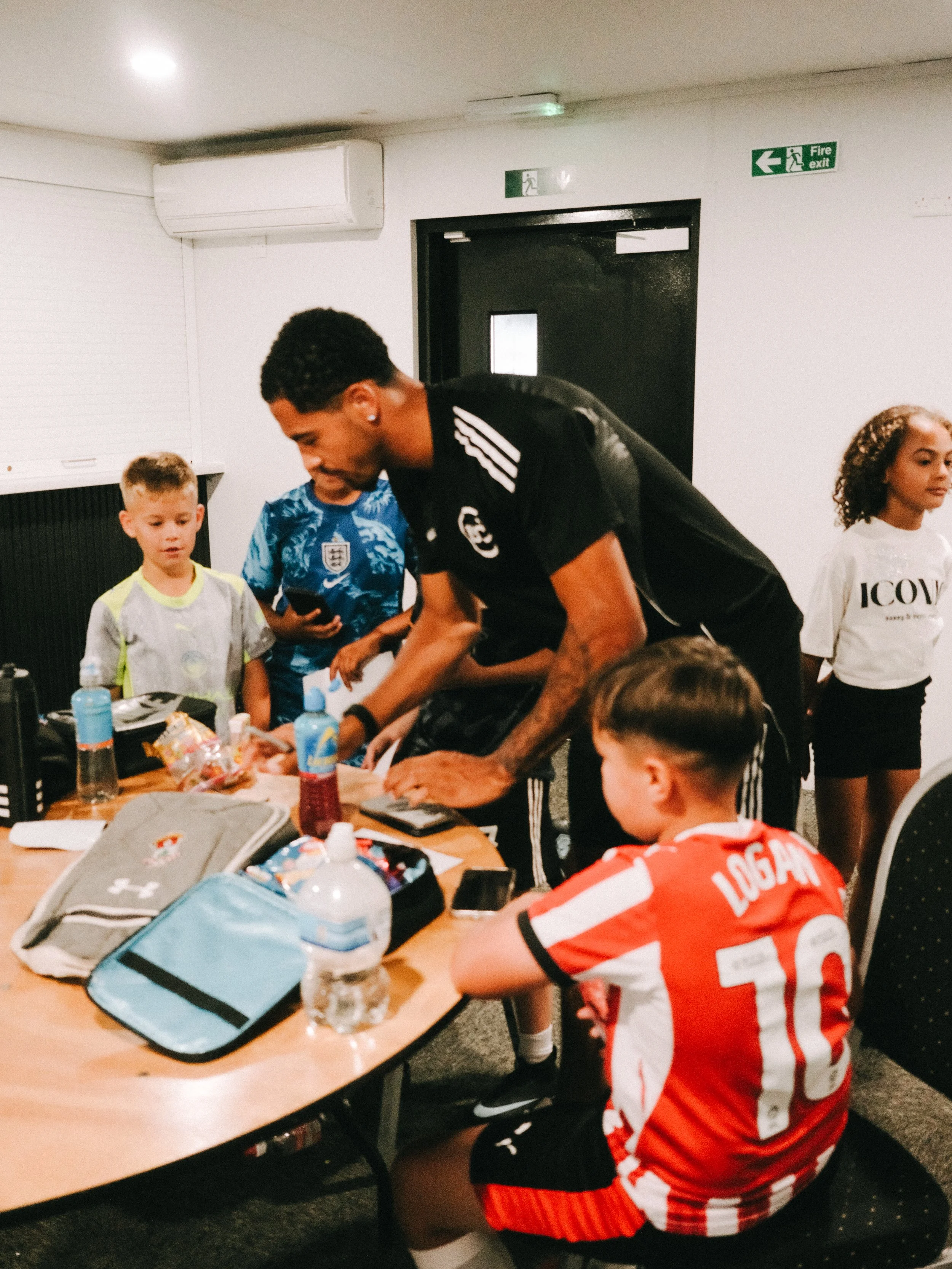 Children and an adult gathered around a table with food and drinks in a room with white walls. Two children are standing, one wearing a gray shirt with green accents and the other in an England football jersey. One boy is sitting at the table, wearin