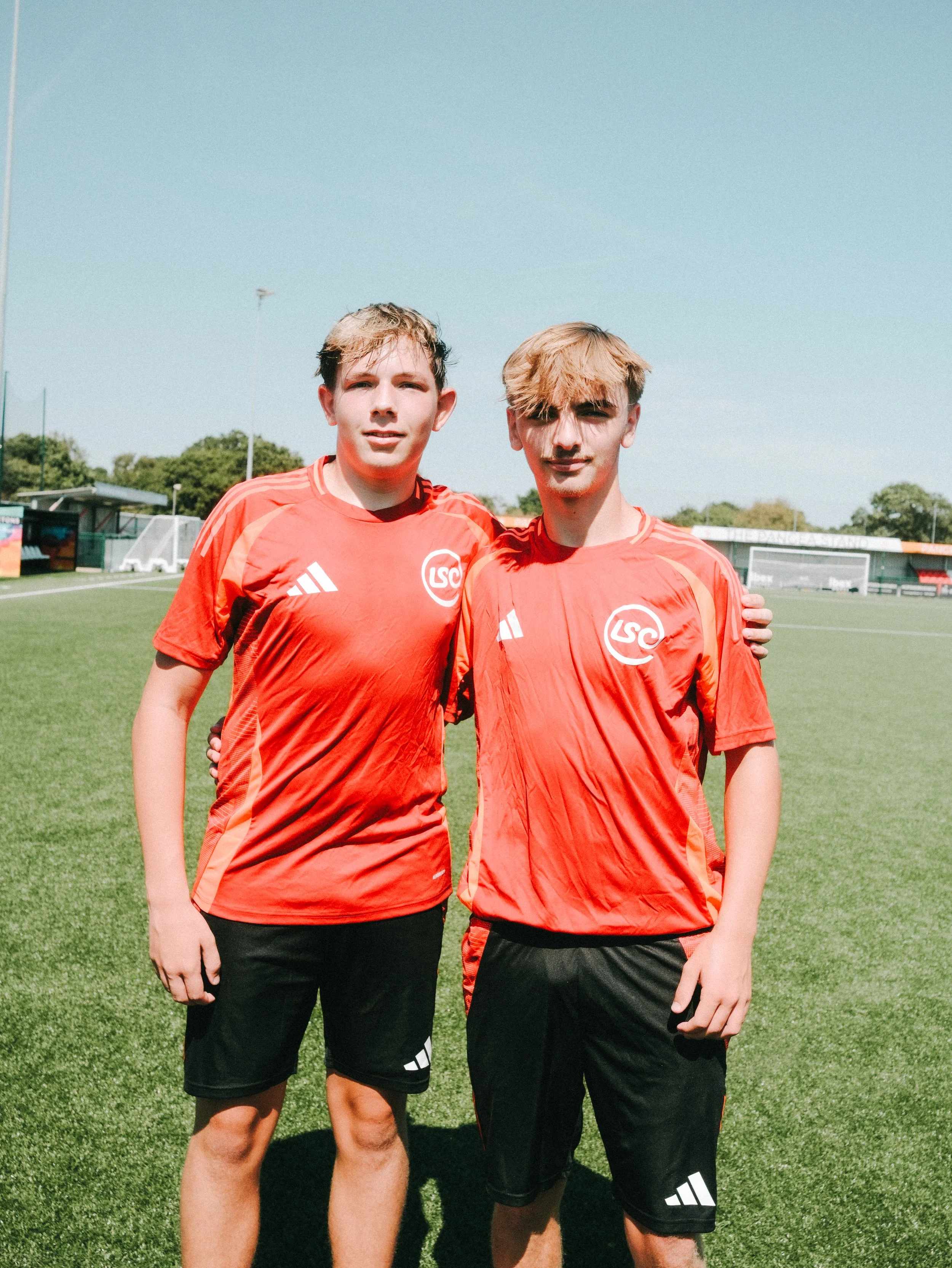 Two young men in red soccer jerseys standing on a soccer field with a stadium and trees in the background on a sunny day.