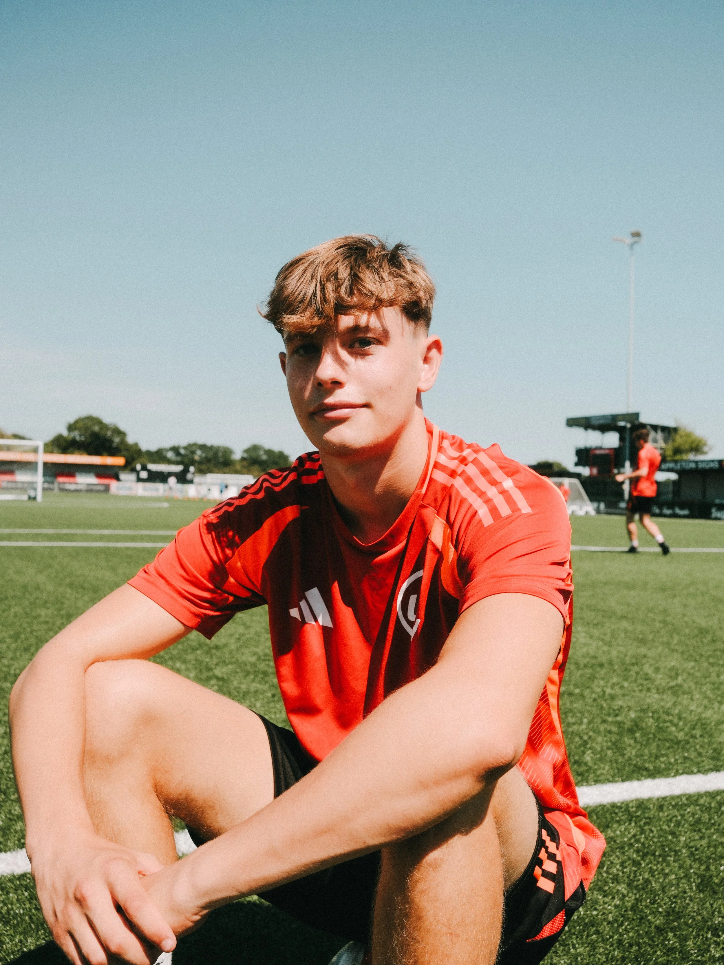 A young male soccer player sitting on the field in a red jersey and black shorts, with a soccer field and players in the background on a sunny day.
