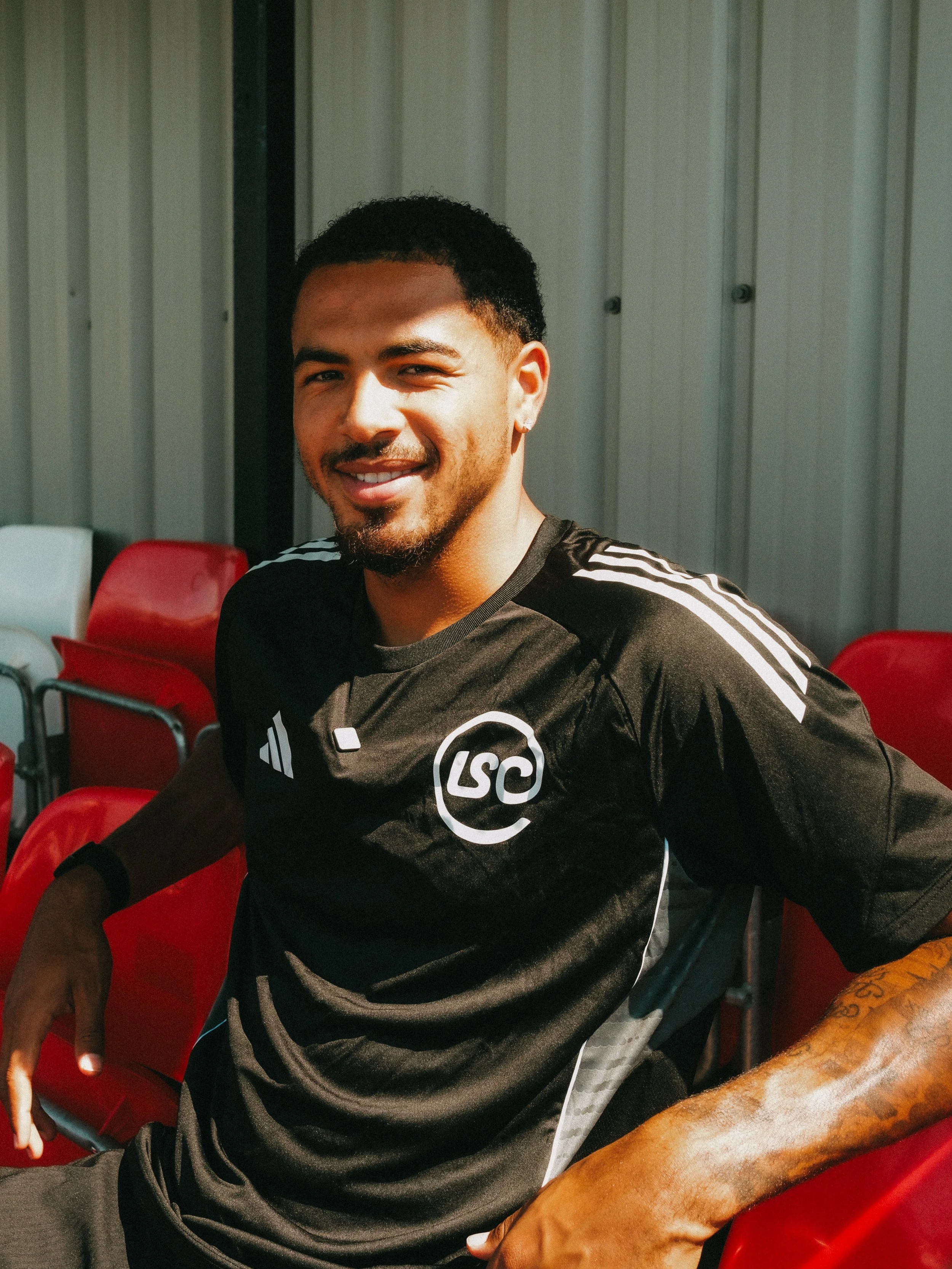 A young man with short dark hair, a beard, and a earring is smiling while sitting in a red chair, wearing a sporty black shirt with white stripes on the shoulders.