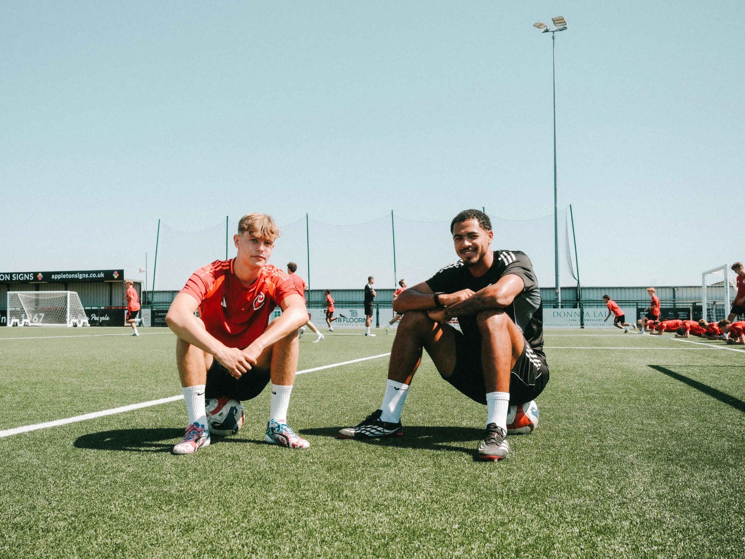 Two young men squatting on a soccer field with the field, goalposts, and other players in the background under a clear blue sky.