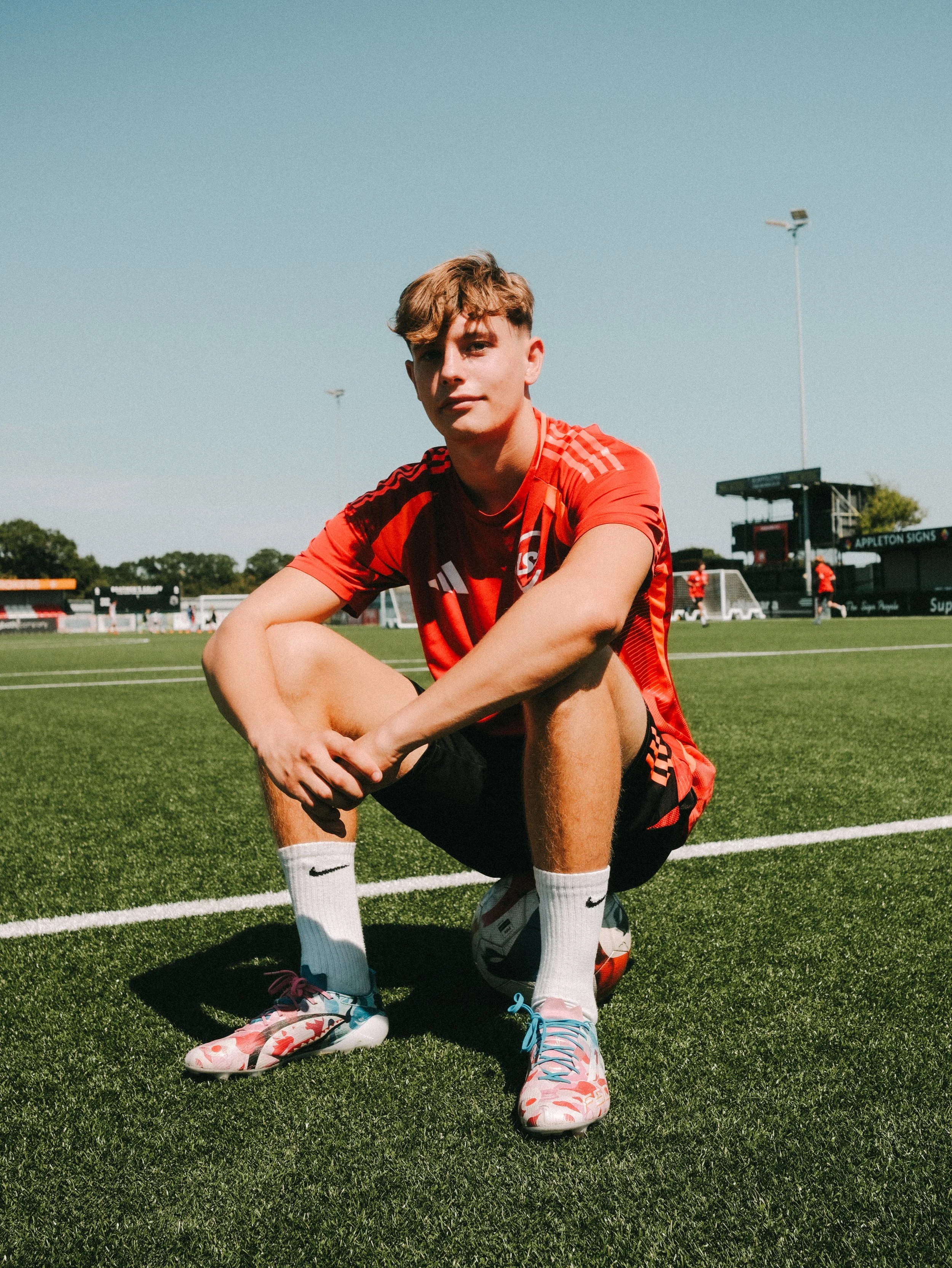 A young male soccer player in a red uniform crouching on a soccer field, with a soccer ball and other players in the background.