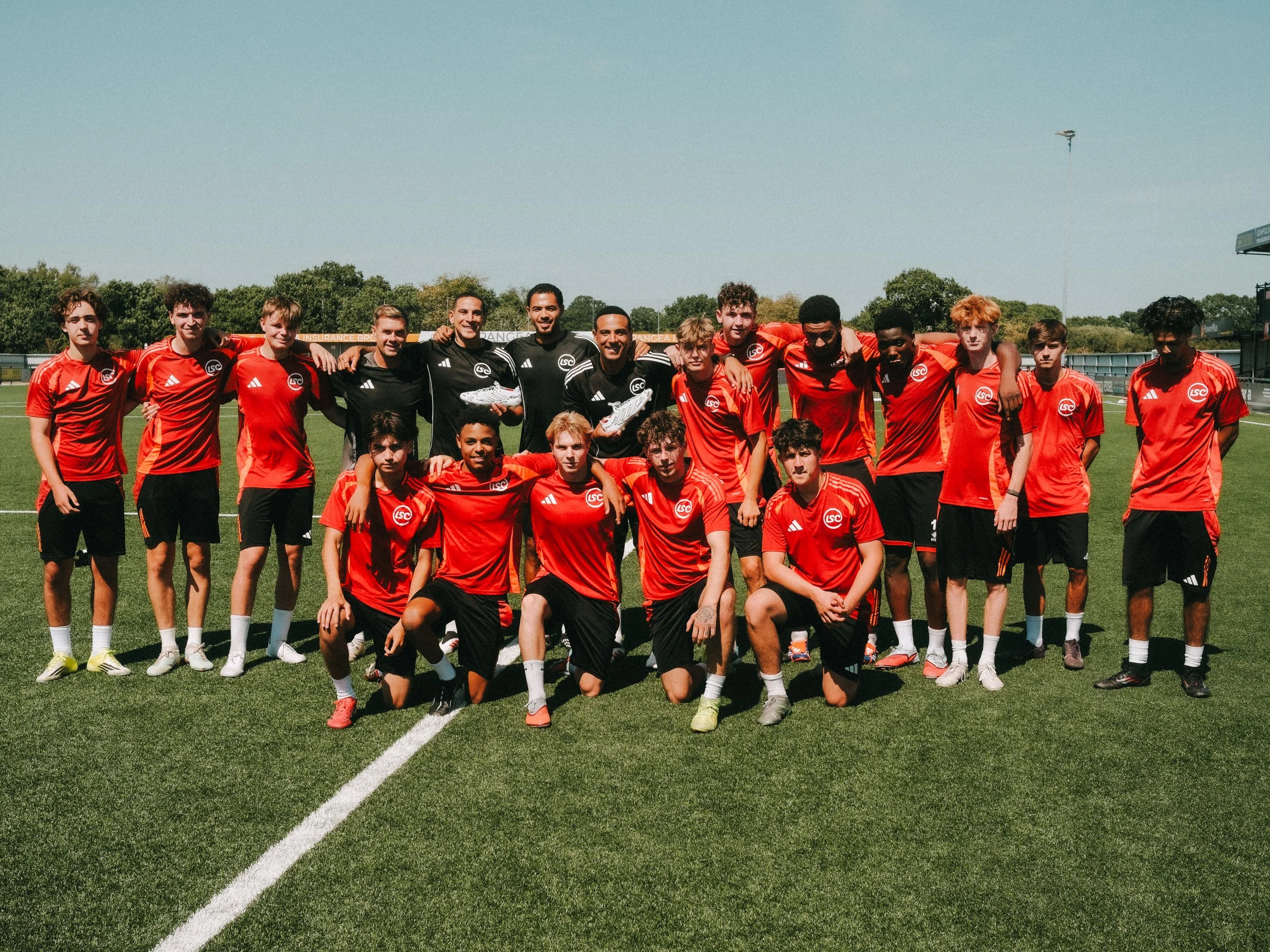 A group of young male football players and coaches standing on a soccer field, posing for a team photo. They are wearing red and black sports uniforms, some holding soccer shoes, with trees and a clear sky in the background.