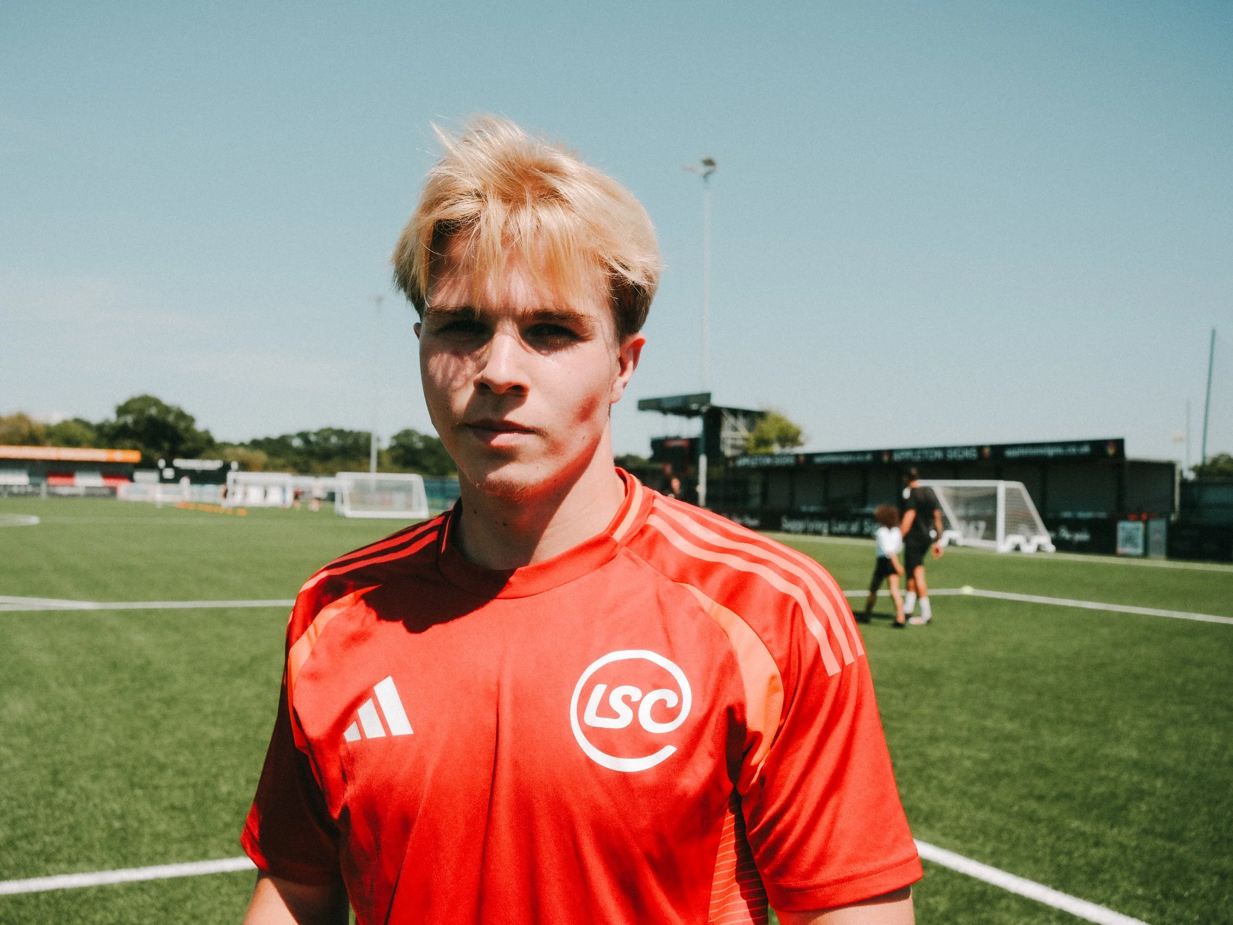 A young male soccer player with blond hair, wearing a red jersey with white logos, standing on a soccer field during daytime.