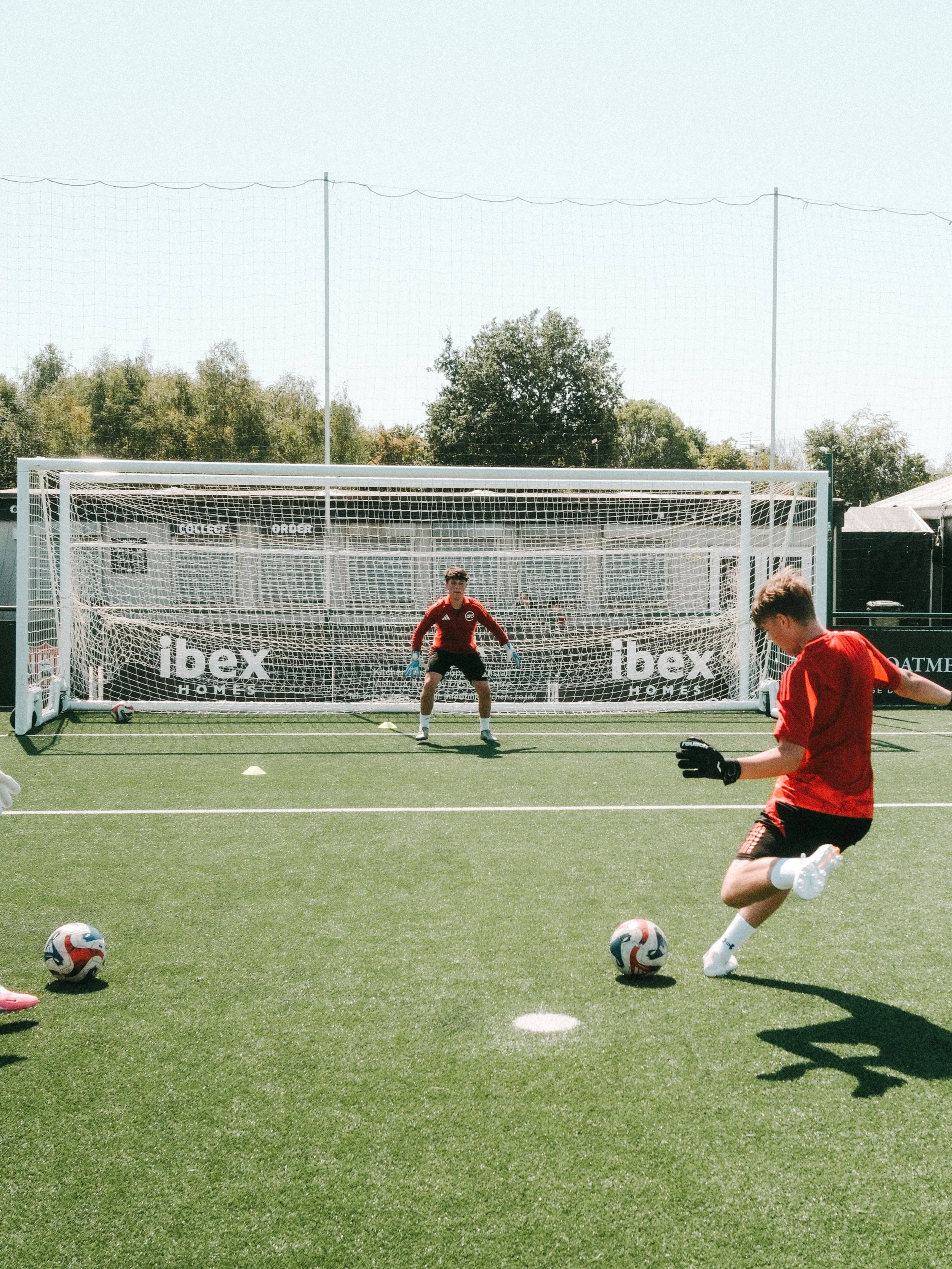 Soccer players practicing on a field with soccer balls, a goalkeeper standing in front of the goal, and a player kicking a ball.