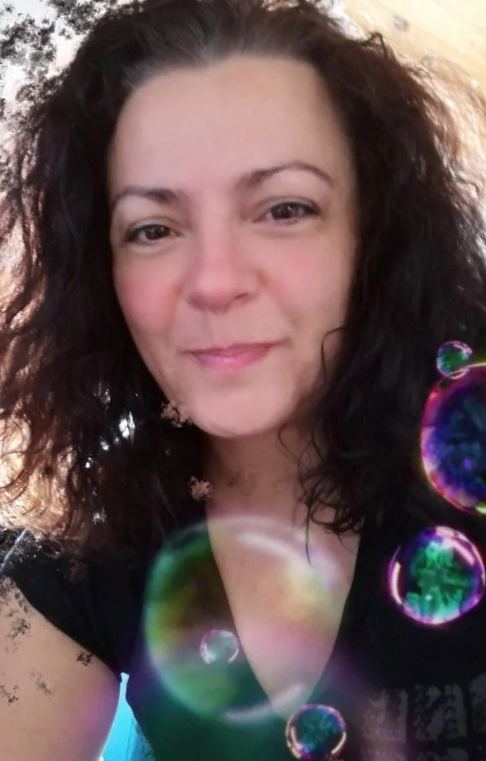 Une femme souriante avec des cheveux bouclés, portant un haut noir, entourée de bulles de savon colorées, en extérieur.