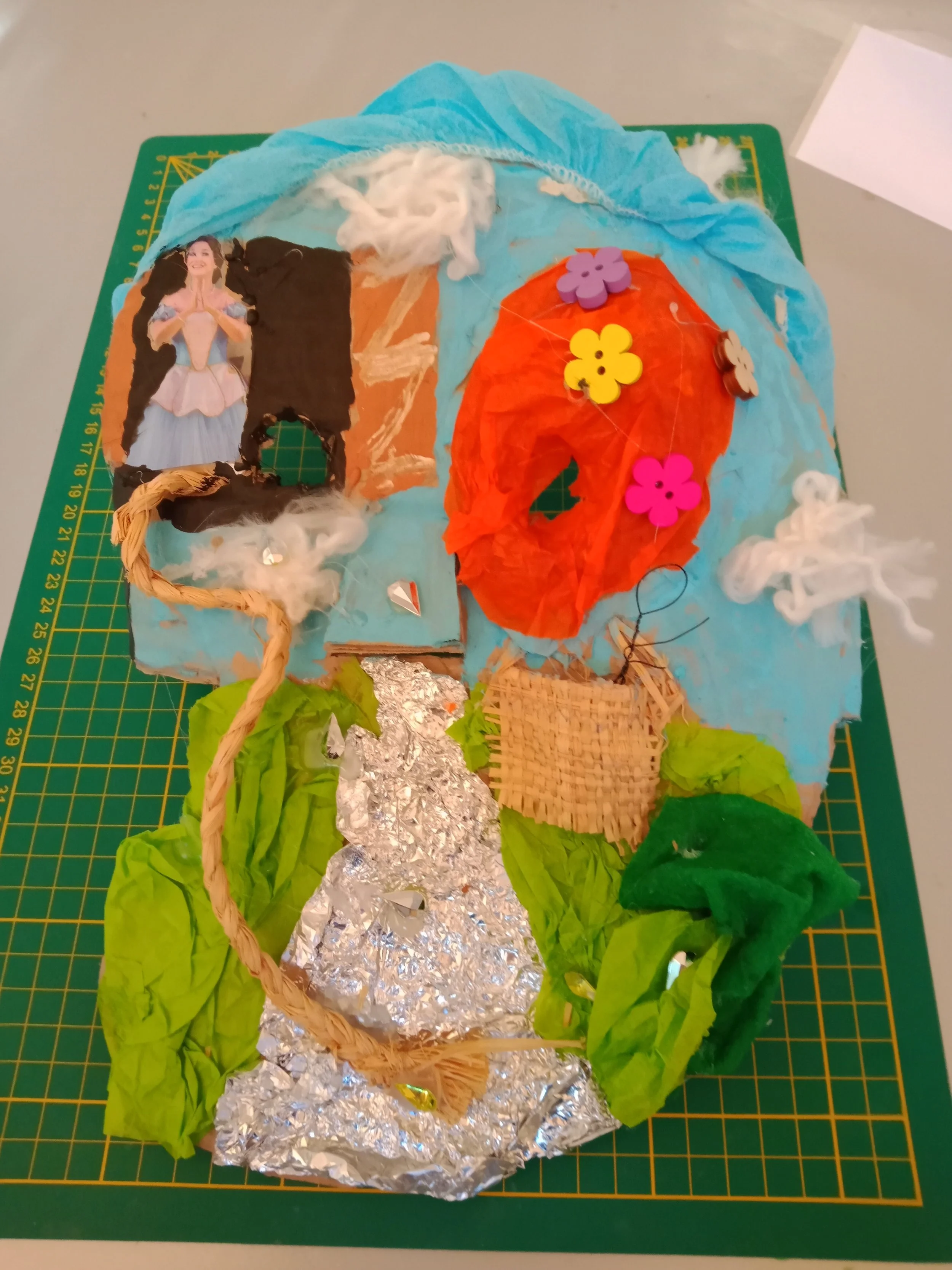 Une création artistique représentant une maison en matériaux recyclés, avec un toit en papier orange, des murs en aluminium, des espaces décorés de fleurs en plastique, des éléments en coton, et des feuillages en papier vert, placée sur une surface de découpe verte.