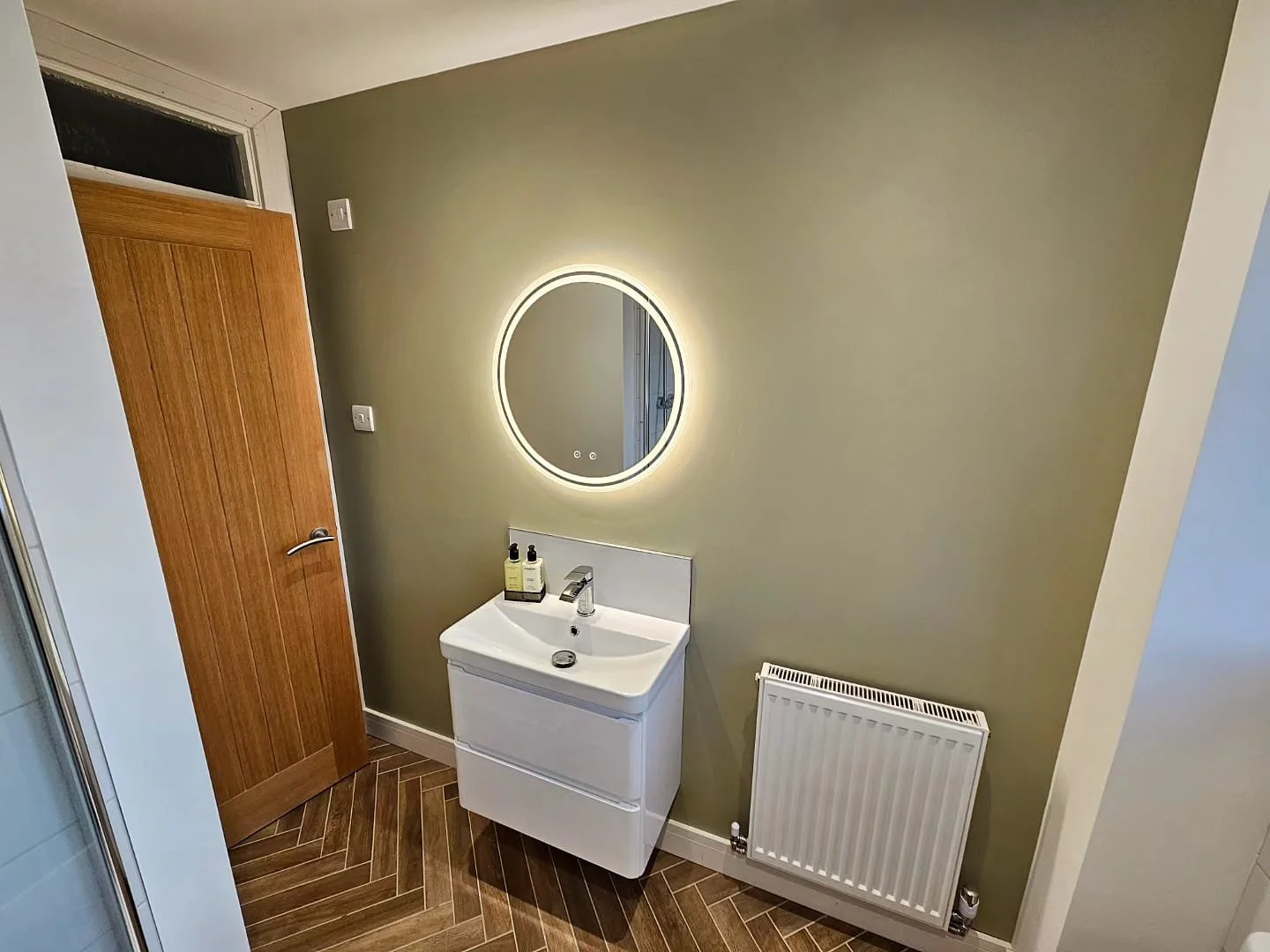 Small modern bathroom with a wooden door, oval LED-lit mirror, white vanity with soap bottles, a radiator, and green walls.
