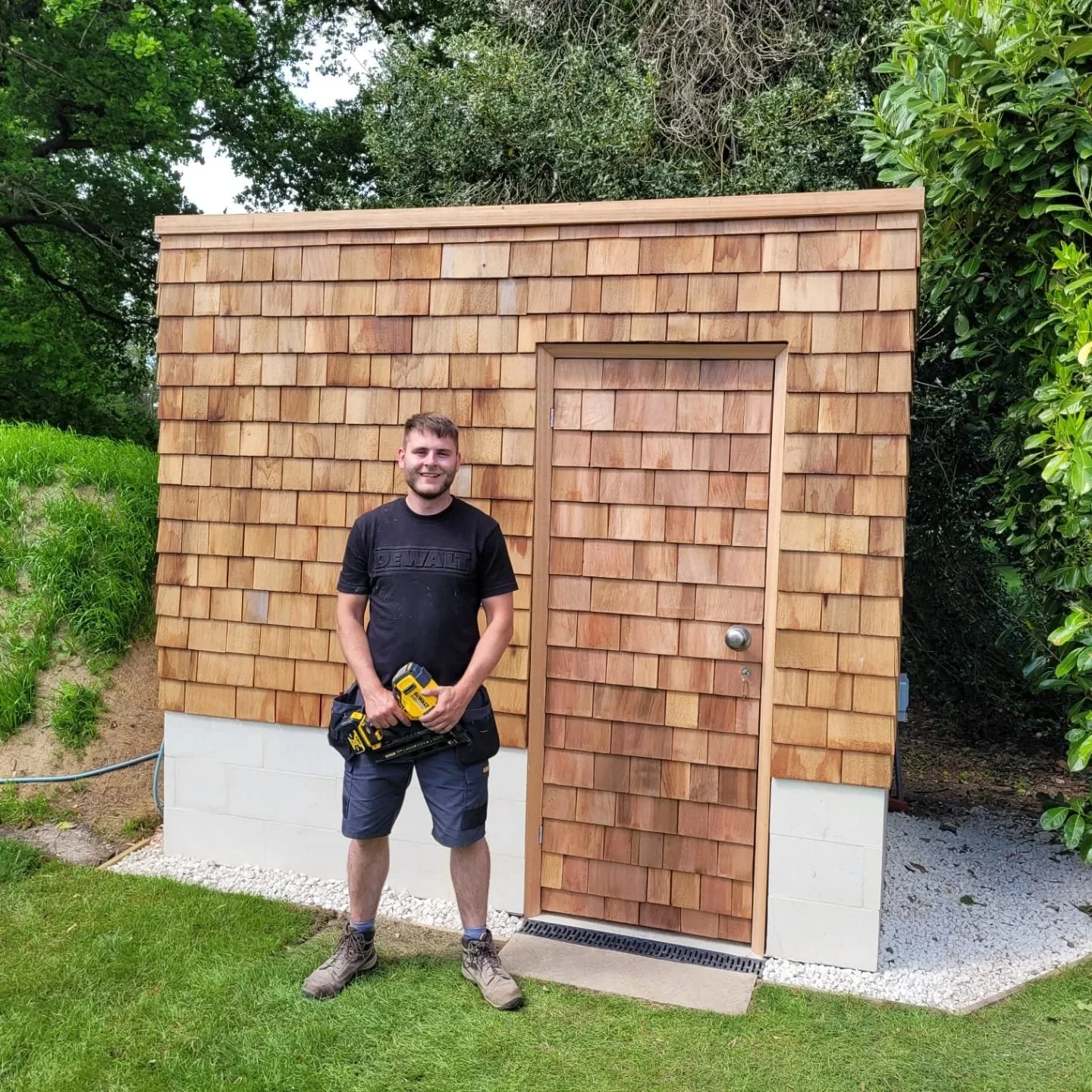 A man standing outdoors in front of a small wooden shed holding a yellow power tool, smiling at the camera, with green trees and grass in the background.