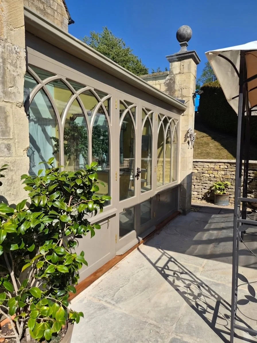 A patio with a glass enclosed sunroom attached to a stone building, a large bush in a pot, and an umbrella with a cast iron table.