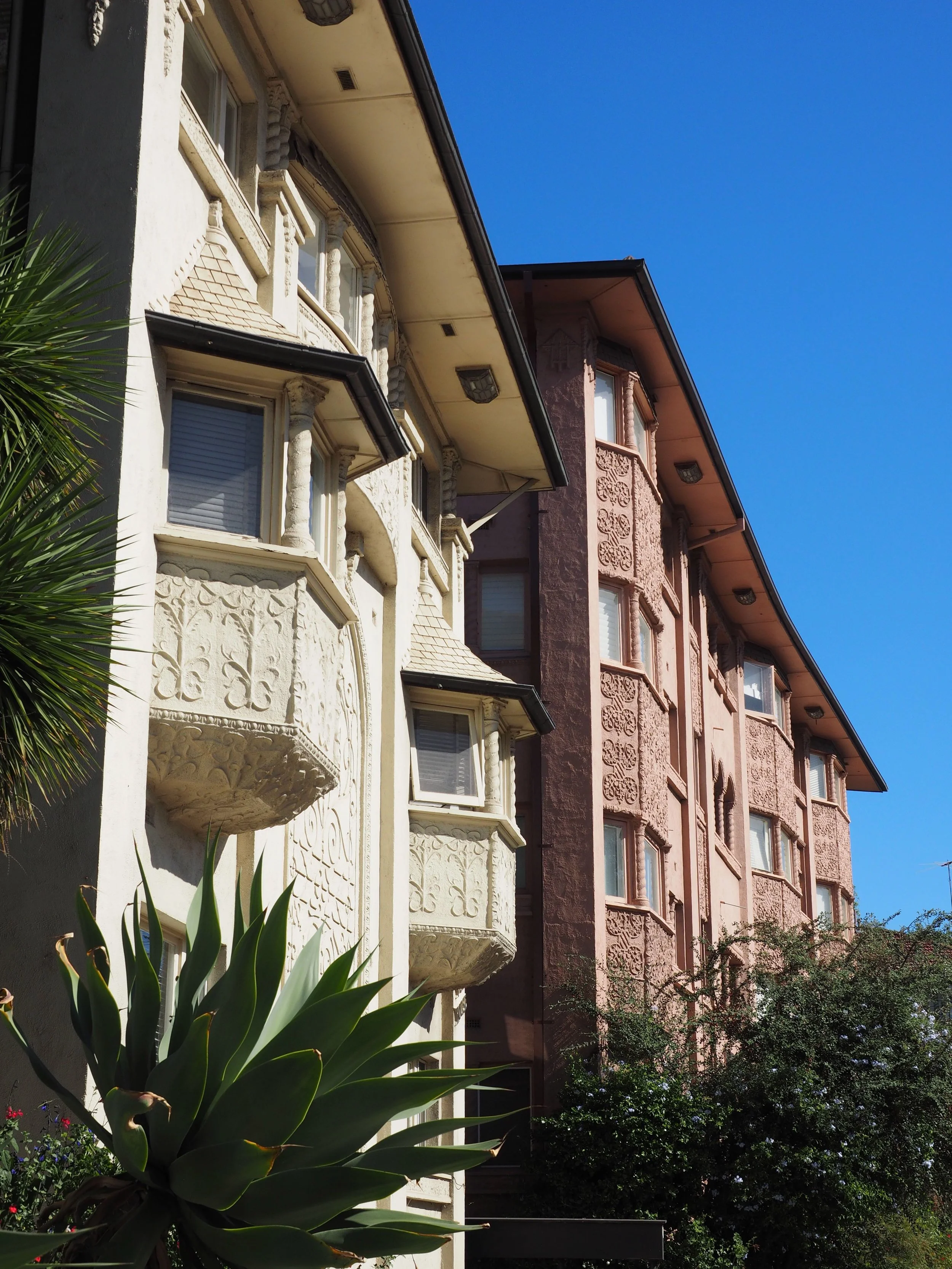 Three ornate multi-story buildings with decorative facades, set against a bright blue sky, with green plants and trees in the foreground.