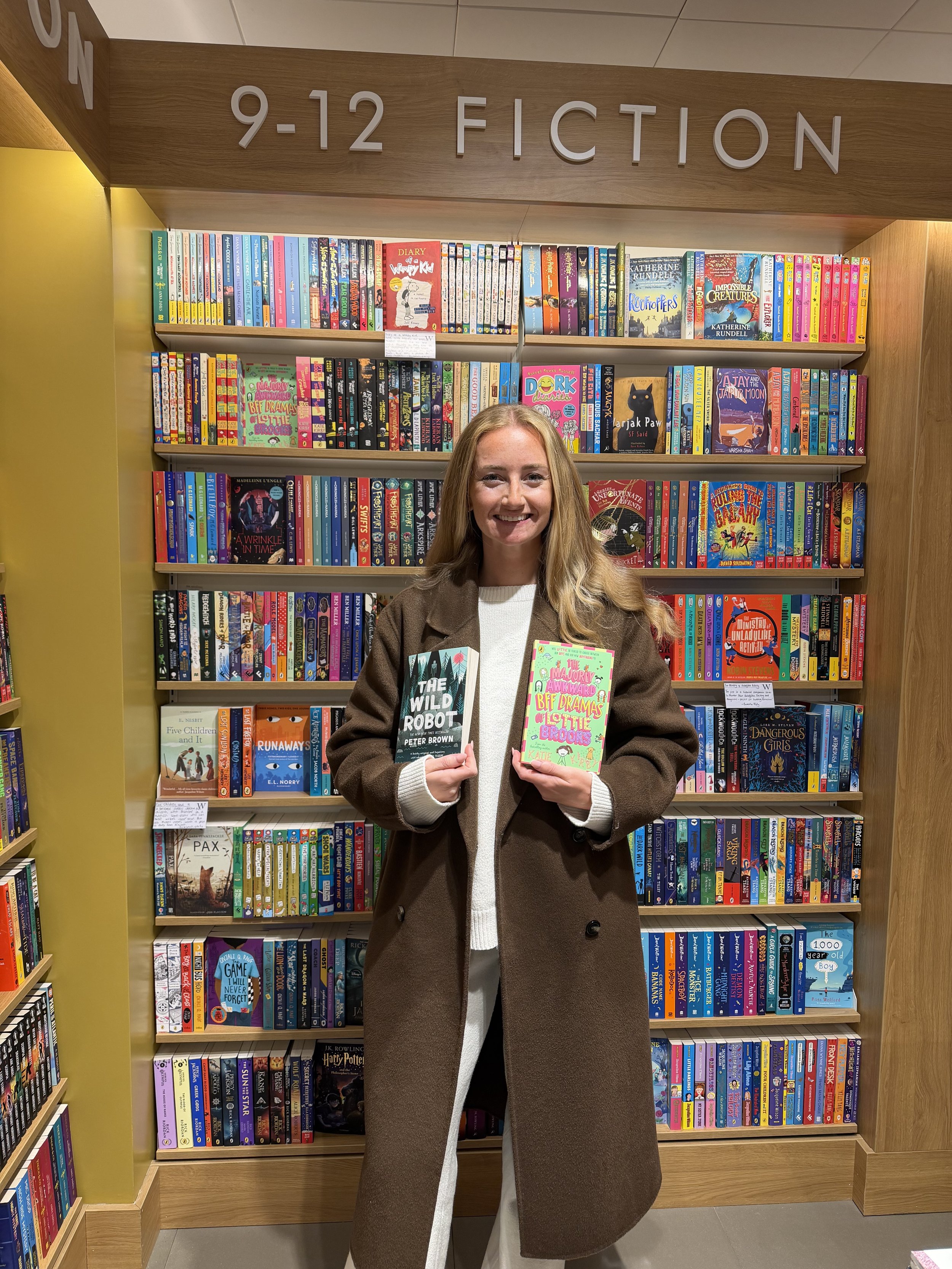 A woman with long blonde hair smiling and holding two books, standing in front of a bookshelf filled with colorful books at a bookstore or library.